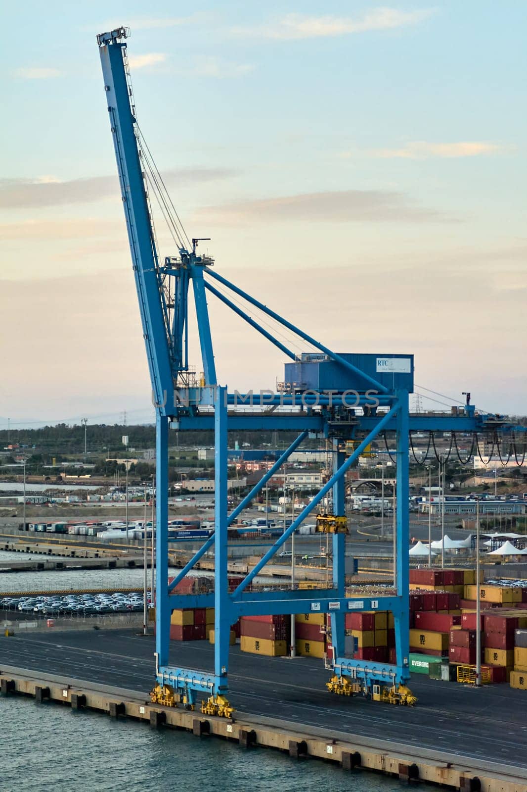 Civitavecchia. Italy - February 17, 2025: A large blue crane at the port of Civitavecchia used for loading and unloading shipping containers. The crane is surrounded by colorful stacked containers.