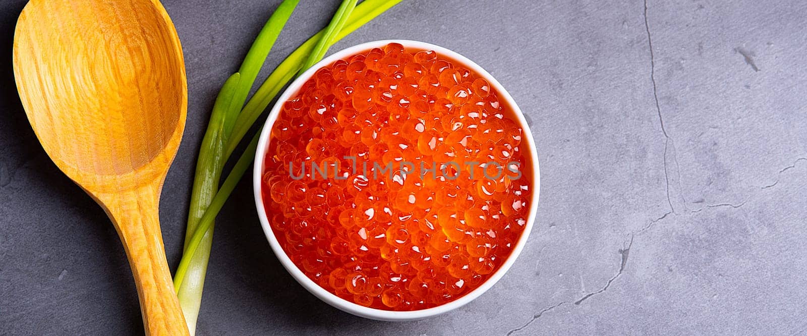 Red caviar in a wooden cup on a gray background with a spoon and green onions. A large pile of bright caviar. Fresh delicious caviar. A place for advertising, logo, label, layout, mockup. Layout, template.