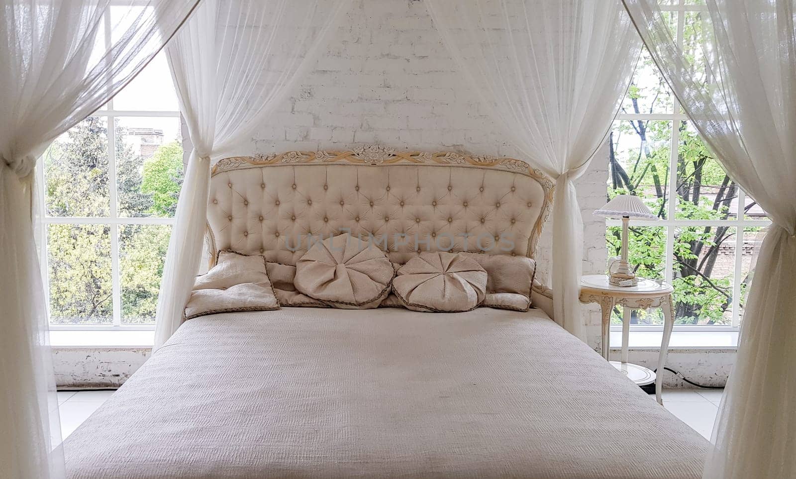 A cozy white wooden bed near a large window with beautiful curtains in the room. Gold-framed modern coffee table with elegant light in a white studio interior with a large bed.