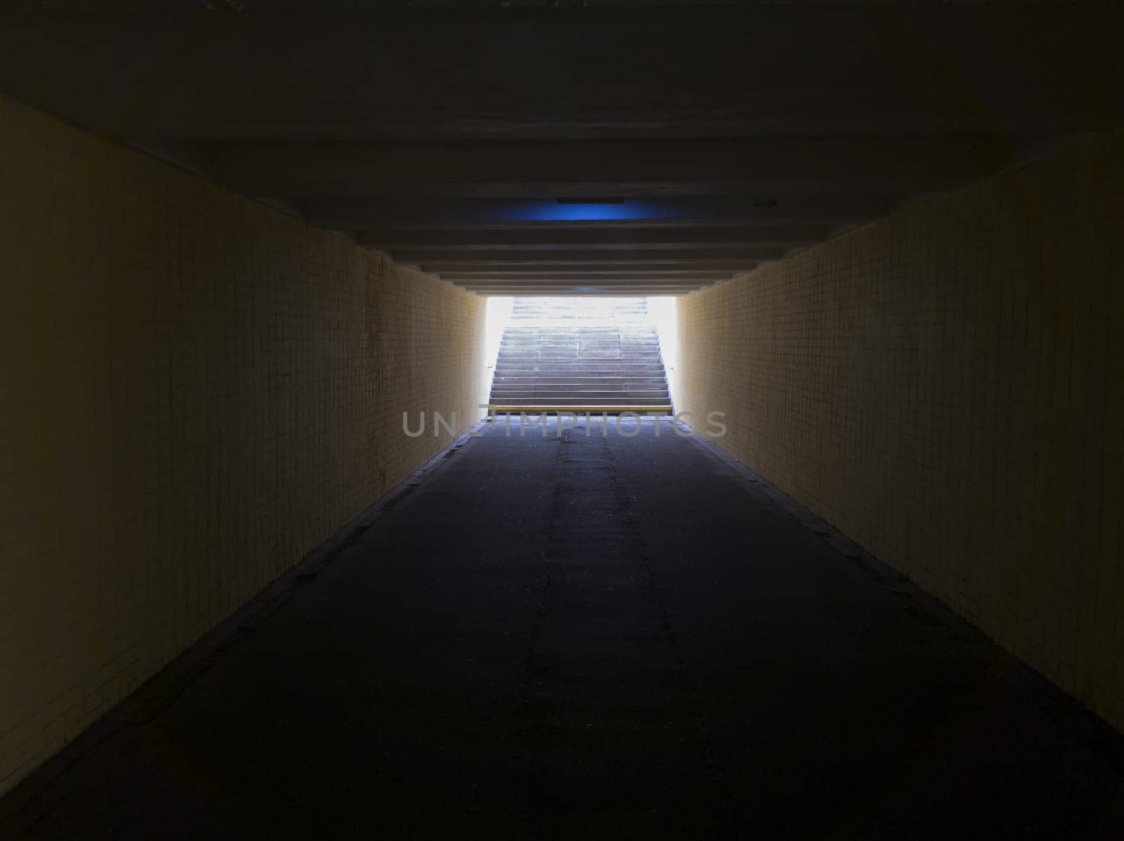Empty yellow underground pedestrian crossing. Tunnel and daylight at the end. Steps to the top at the pedestrian crossing. A long concrete tunnel with lanterns in the city underground