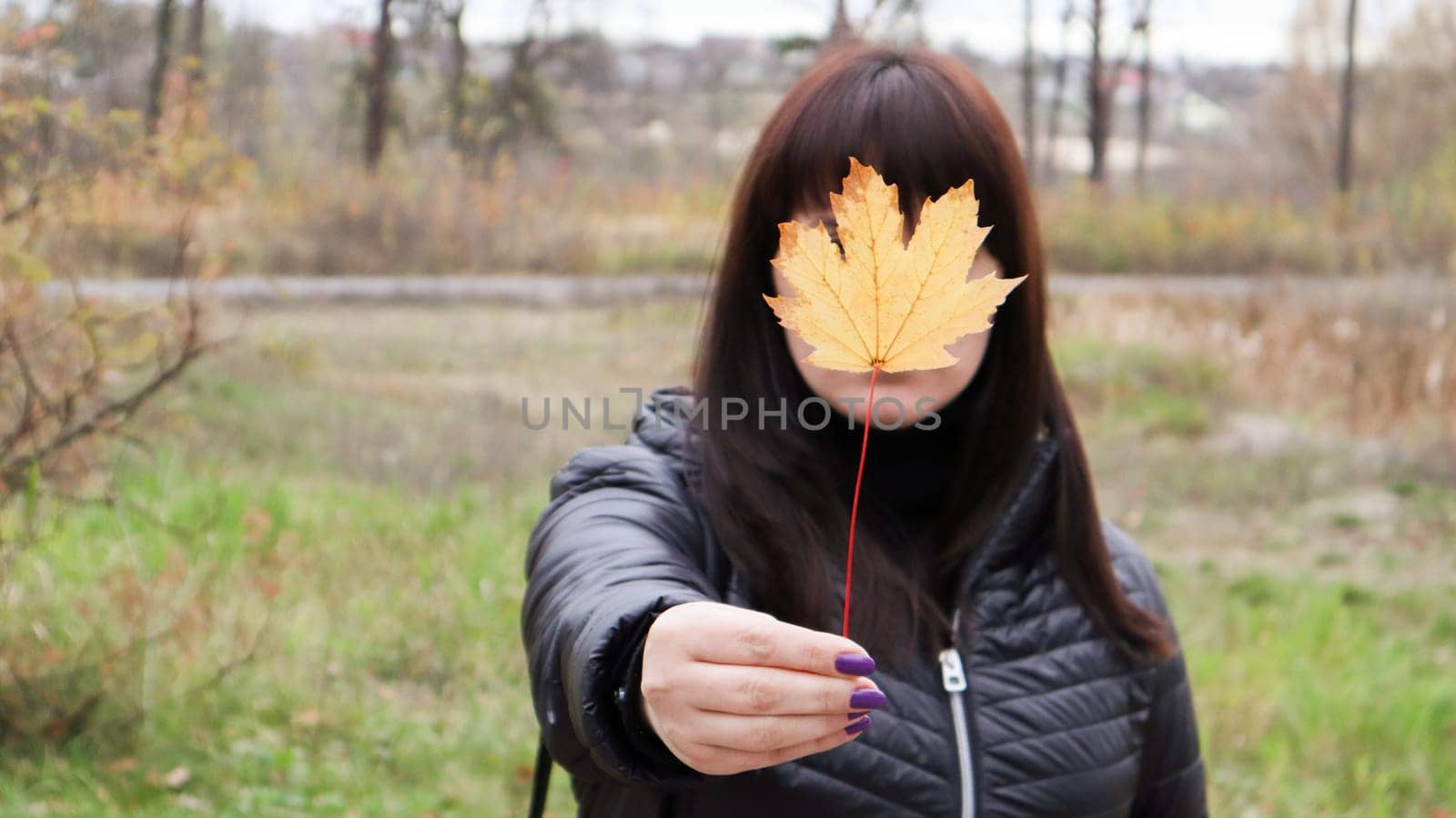 Girl holds a yellow maple leaf in front of her. A young woman covered her face with a yellow autumn maple leaf in a park. Lovely girl enjoying the warm weather. Autumn mood. Enjoy the season.