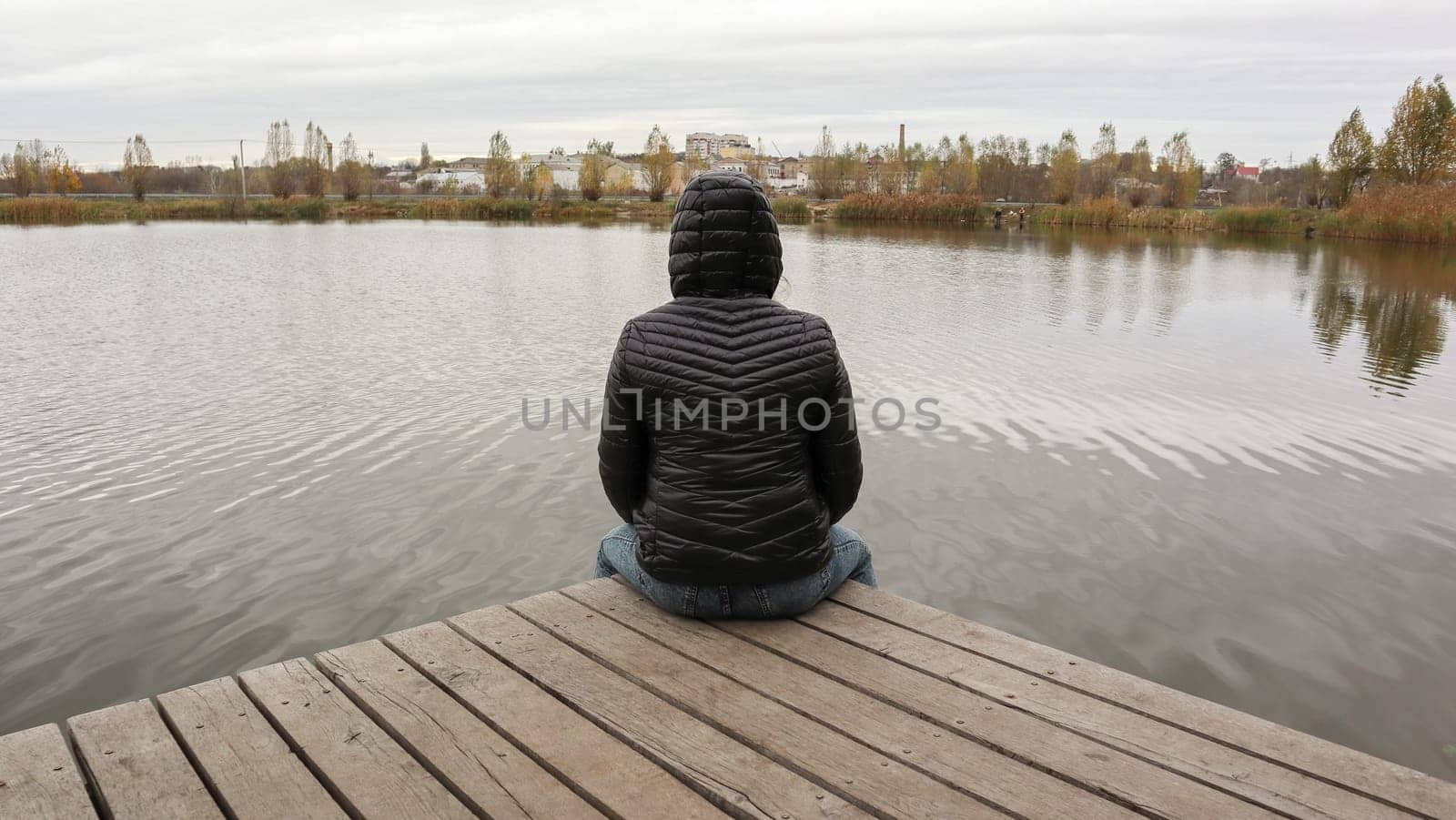 A girl in a jacket sits on a wooden bridge by the lake in cold autumn weather. Dipresia or dreaming alone. Thinking one on one with nature