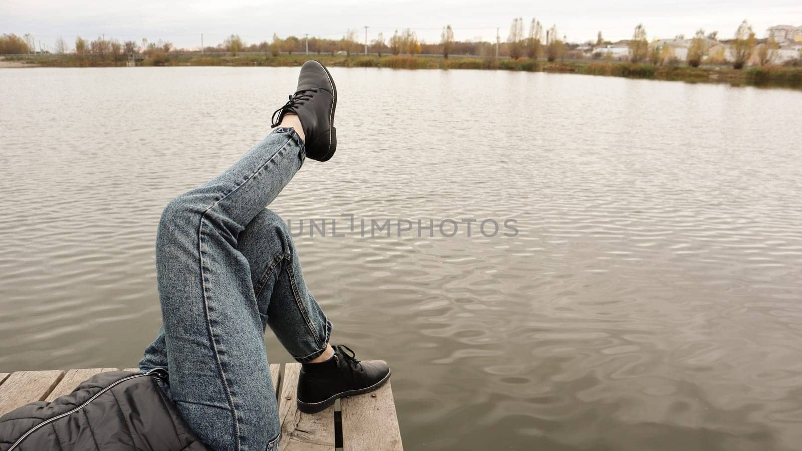 A girl in blue jeans sits by the lake. A young woman is resting by the river, sitting on the edge of a wooden bridge. Pair of legs relaxing by a lake on a wooden dock. People are resting.