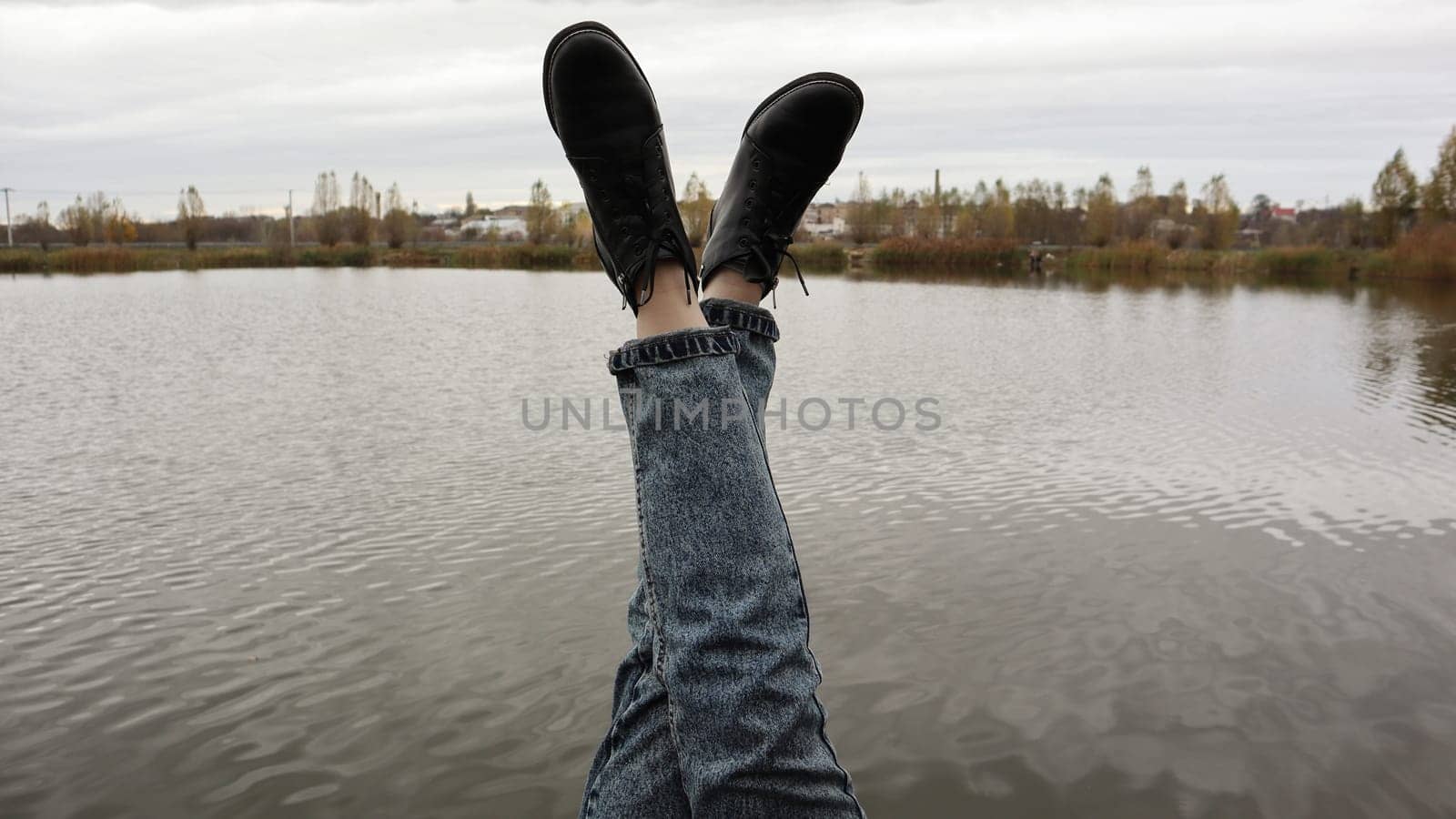A girl in blue jeans sits by the lake. A young woman is resting by the river, sitting on the edge of a wooden bridge. Pair of legs relaxing by a lake on a wooden dock. People are resting