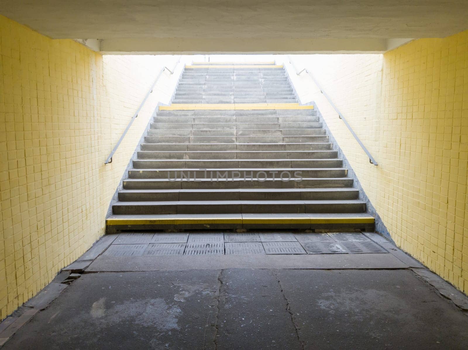 Empty yellow underground pedestrian crossing. Tunnel and daylight at the end. Steps to the top at the pedestrian crossing. A long concrete tunnel with lanterns in the city underground