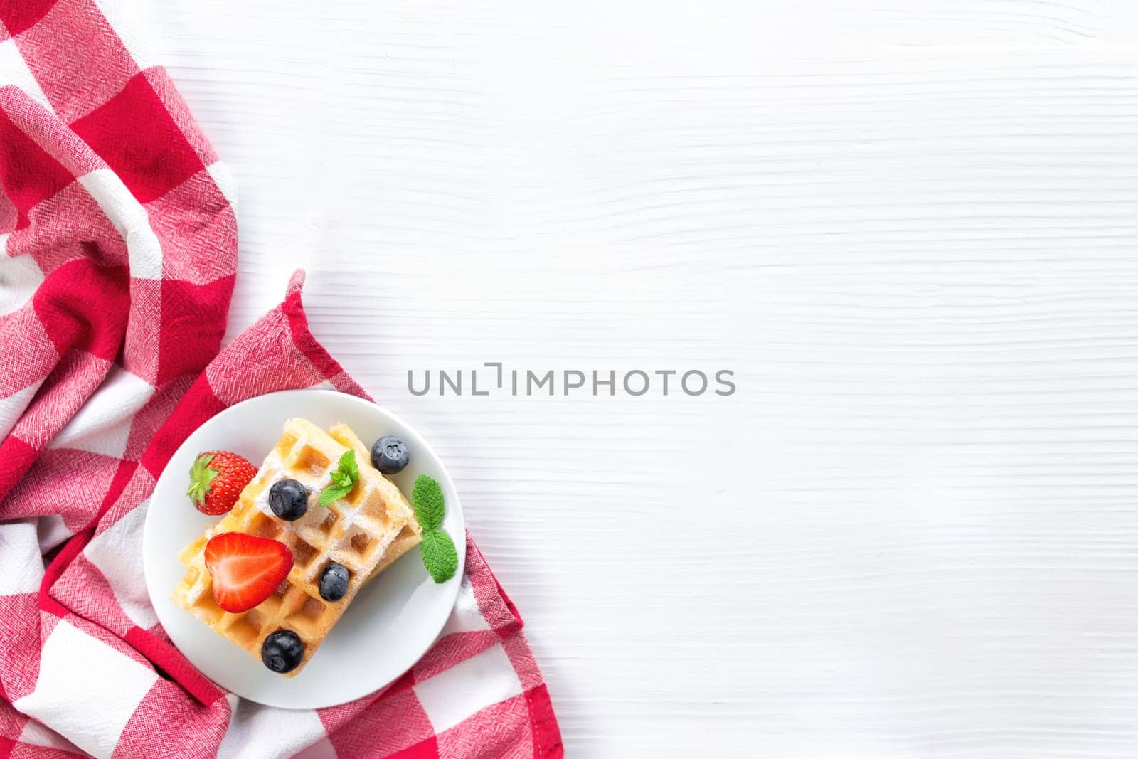 White plate on red kitchen towel with stack homemade Belgian waffles, blueberry, strawberry cuts, mint leaf, sugar powder on gray background