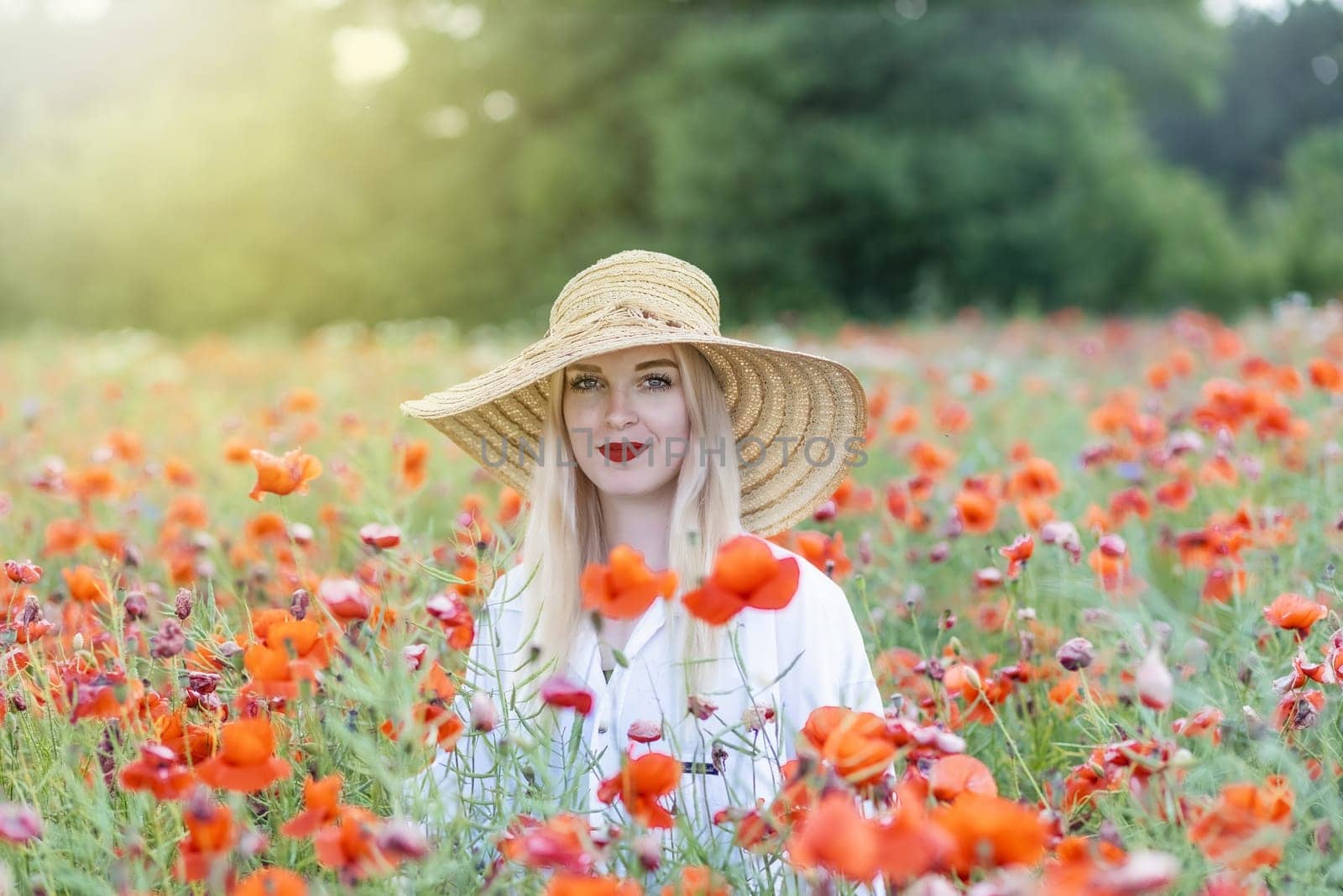 Portrait of blonde young woman in straw hat posing in red poppy field smiling at the camera. Horizontally.