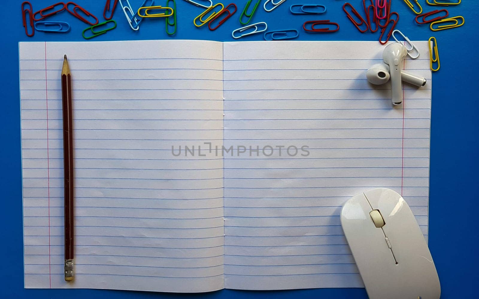 Flat lay photo of a school desk with a mouse, blank open notebook with pencil, paper clips, earphones on a blue wooden tabletop.