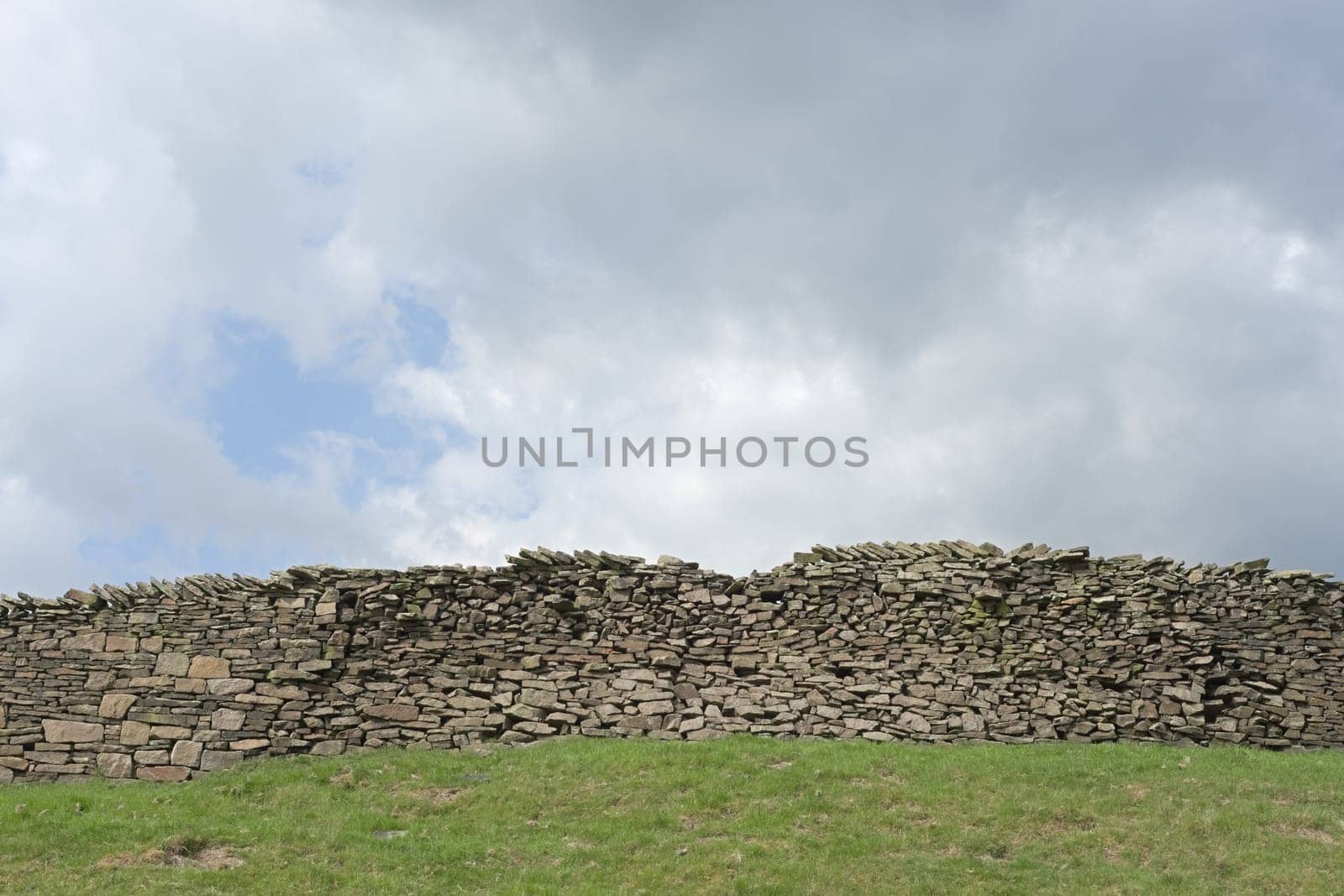 Large dry stone wall and a heavily clouded blue sky background. Agricultural and rural theme, green grassy field foreground. Plenty of copy space.