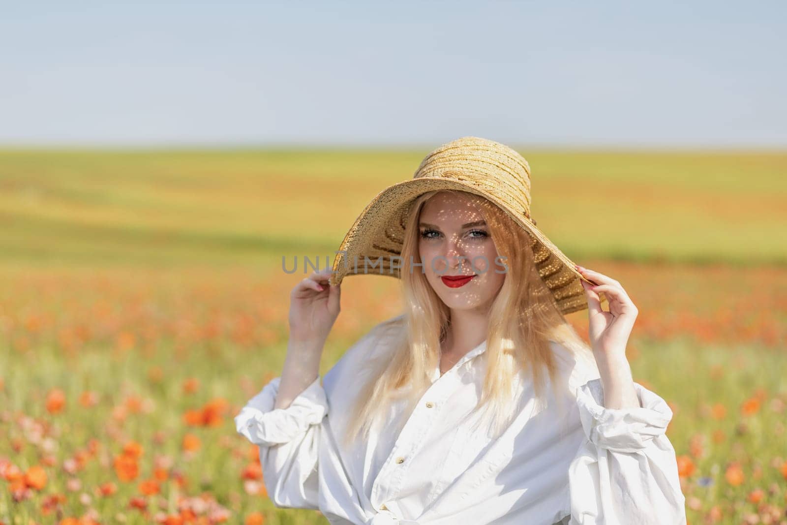 Attractive young woman is standing n a field of red poppies holding her straw hat on the head. Horizontally.