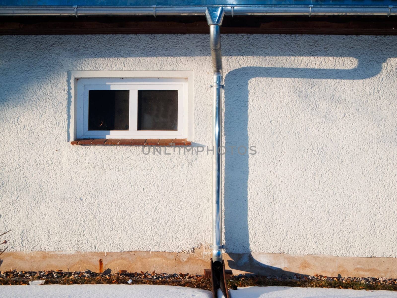 Detailansicht einer flachen Hausfassade mit Dachrinne, Fallrohr und kleines Fenster im seitlichen Abendlicht mit Schattenbild des Fallrohrs.
