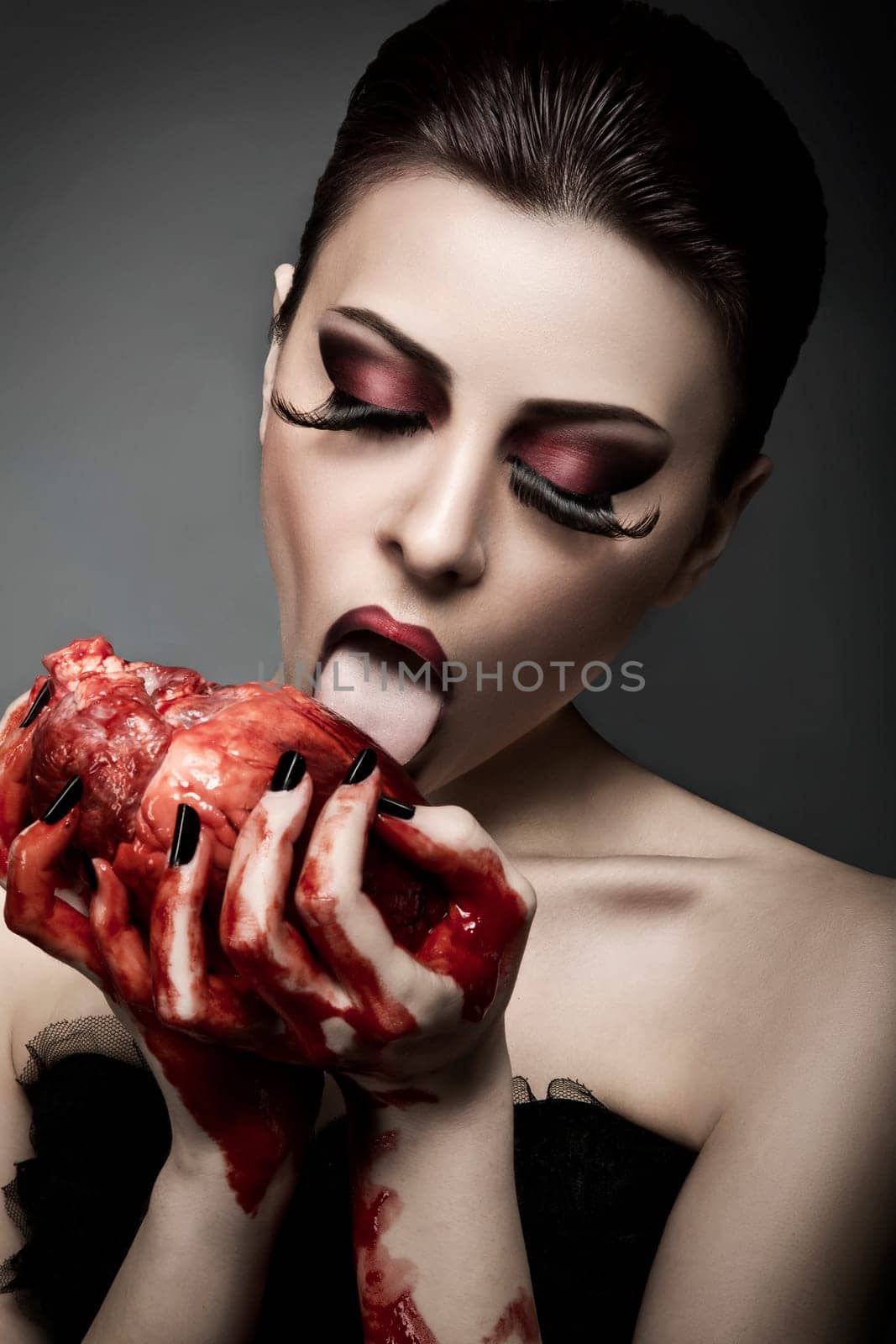 Beauty young woman licks blood from human heart against grey background