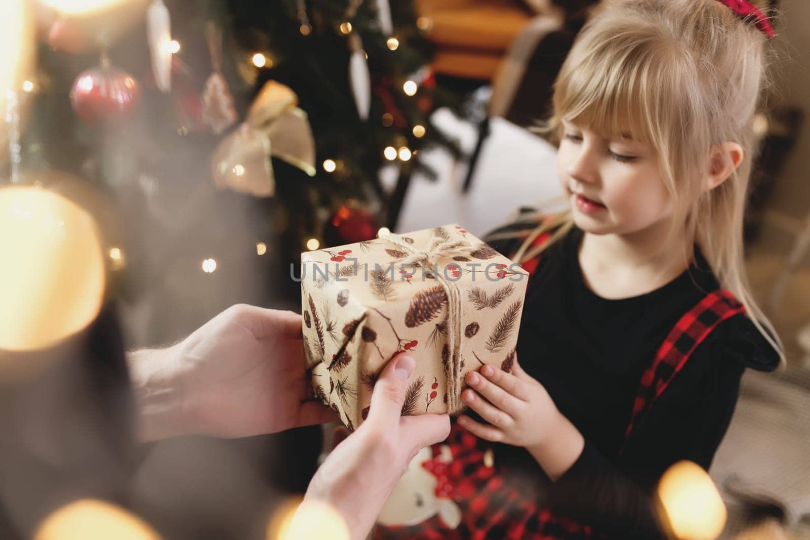 Young father in red Santa hat giving Christmas gift to his smiling daughter near decorated Christmas tree. Girl dressed in festive red-black Christmas outfit. They smiling, happy because of New Year