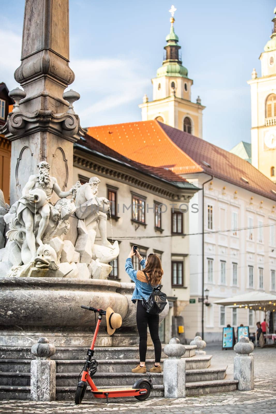 Traveler exploring old town on environmentally friendly electric scooter. Female tourist exploring Ljubljana's old medieval historical city center, taking picture of a fountain with her phone
