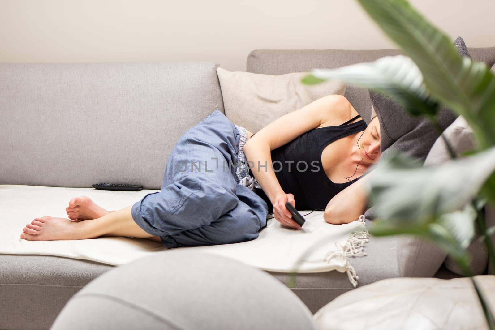 Tired young woman, resting, lying on a comfortable sofa with big cushions in home living room using her mobile phone
