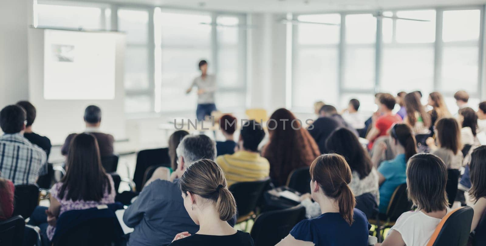 Business and entrepreneurship symposium. Speaker giving a talk at business meeting. Audience in conference hall. Rear view of unrecognized participant in audience.