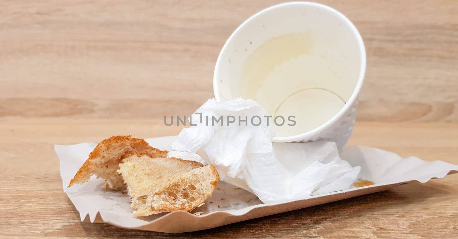 Leftover food on a paper plate and empty dirty paper coffee cup on a wooden table in a fast food cafe.