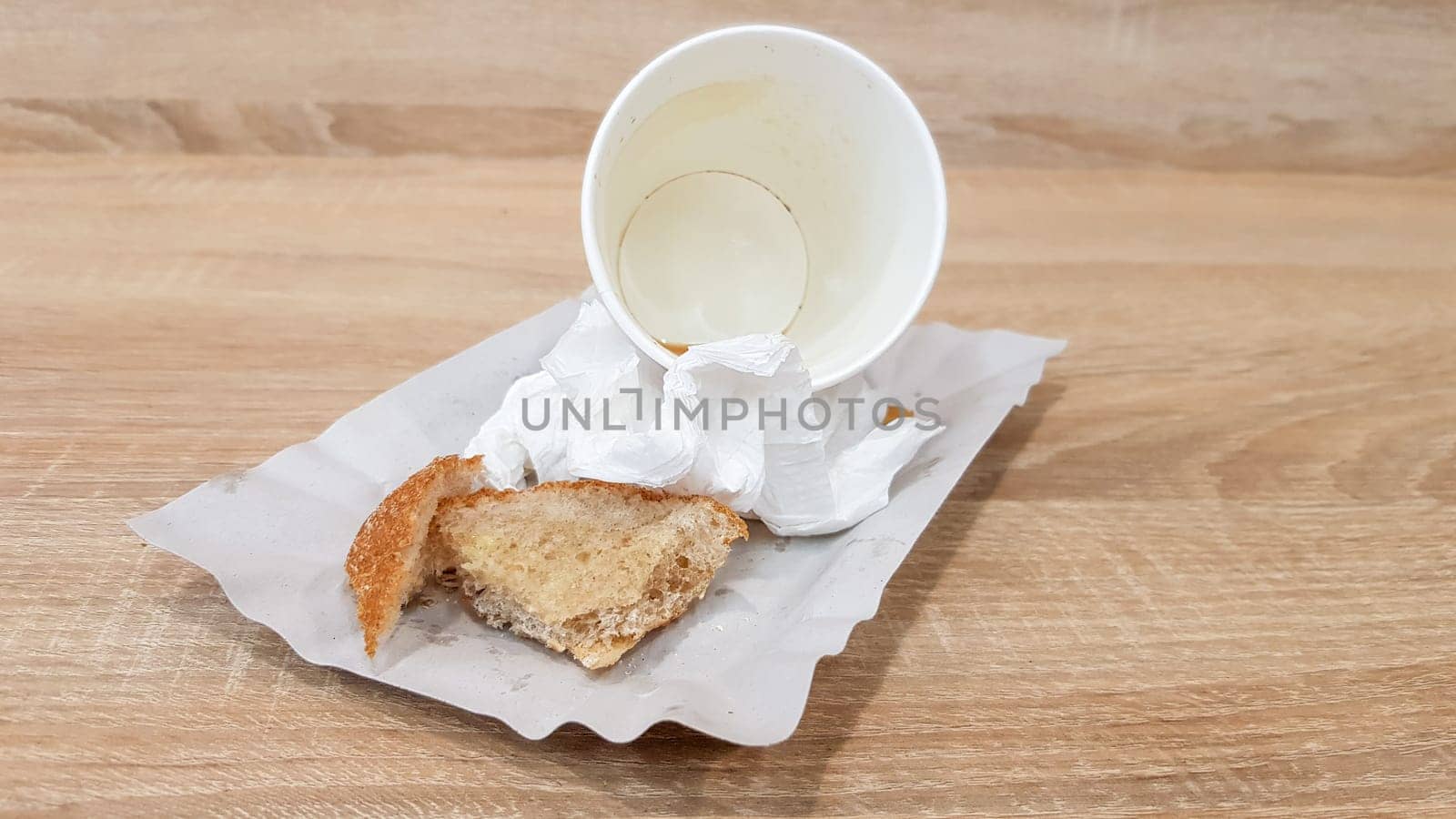 Leftover food on a paper plate and empty dirty paper coffee cup on a wooden table in a fast food cafe.