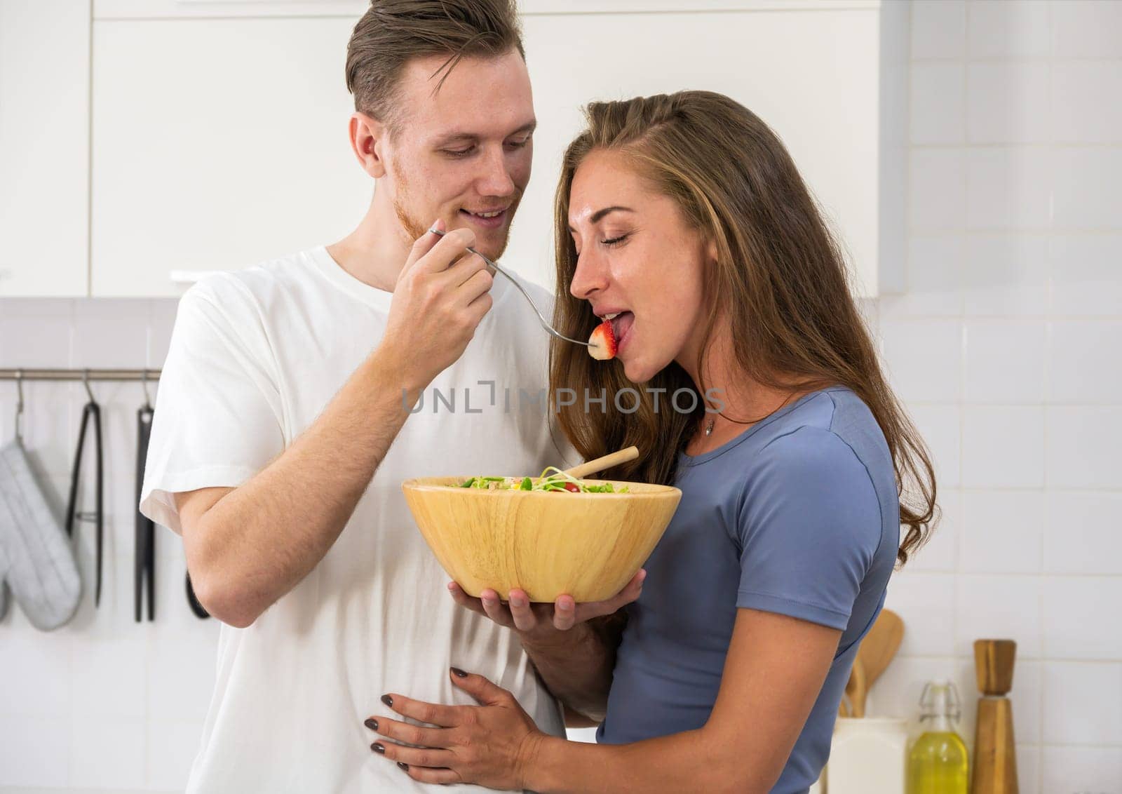 Happy young couple enjoying and preparing a healthy meal in home kitchen. Man holding salad bowl and offering a strawberry to woman