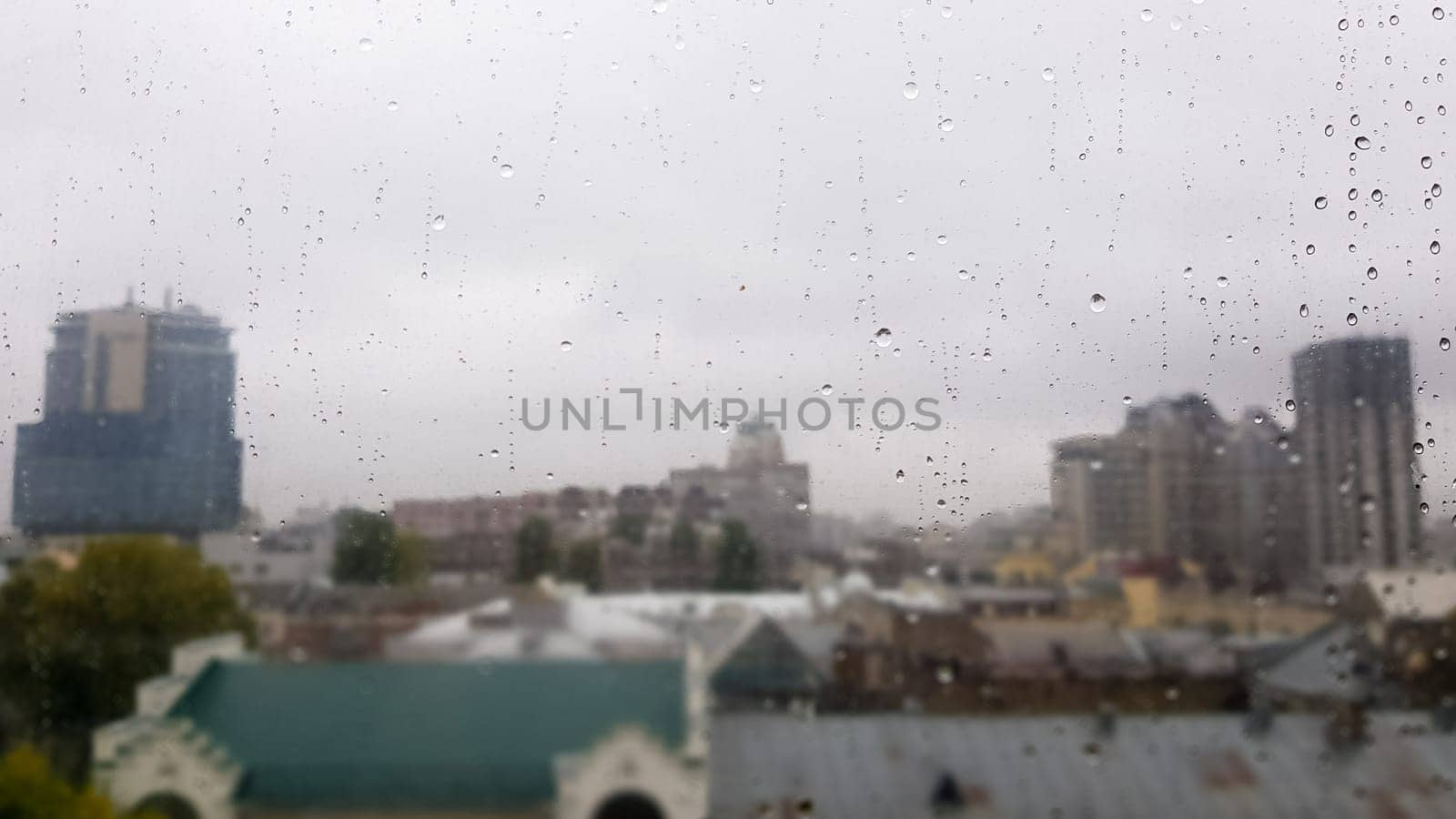 Wet window with drops on the background of the autumn city in cloudy weather. View from the window in the rain. Water drop on glass window during rain with blurred background of city scene.