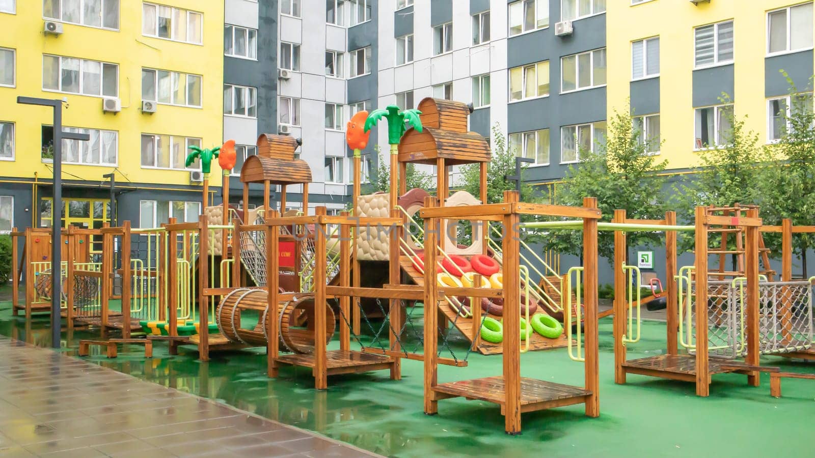 A courtyard of high-rise buildings with a modern and large playground made of wood and plastic on a rainy summer day without people. Empty outdoor playground. A place for children's games and sports