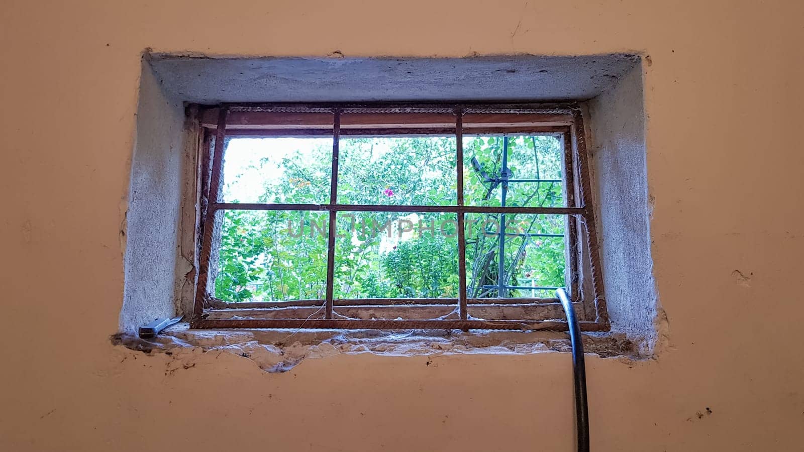 A rectangular basement window is tightened with a metal mesh and a grill. Technical window in the wall of an old gray building