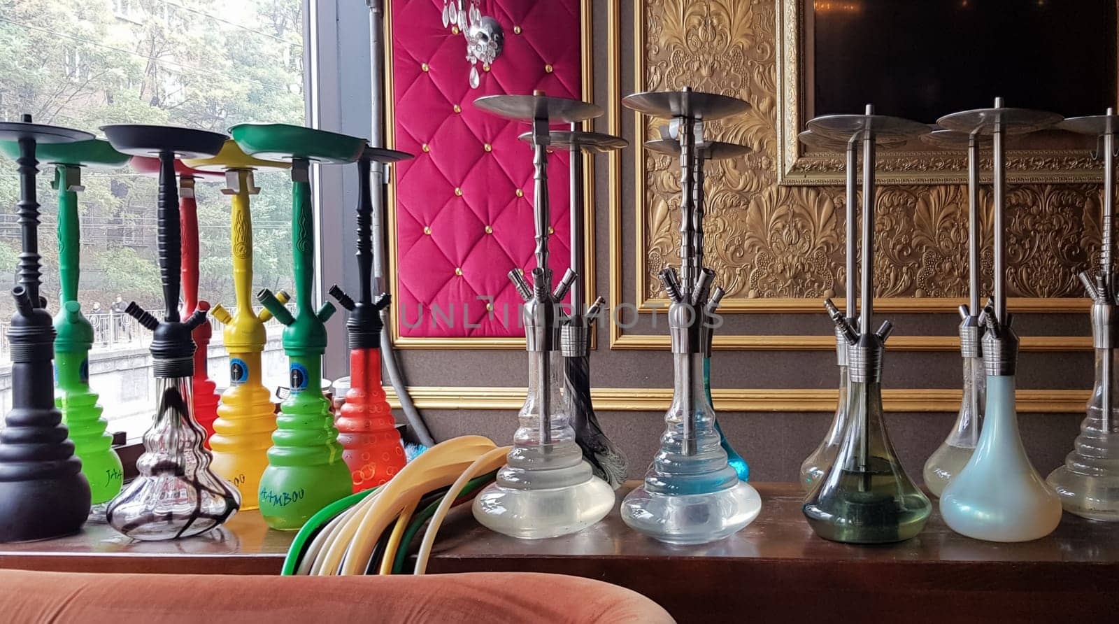 Group of modern glass hookah appliance on a wooden stand. Restaurant bar. Glass hookahs on the bar in a cafe or bar made of beautiful multi-colored glass. Diffused daylight from the window