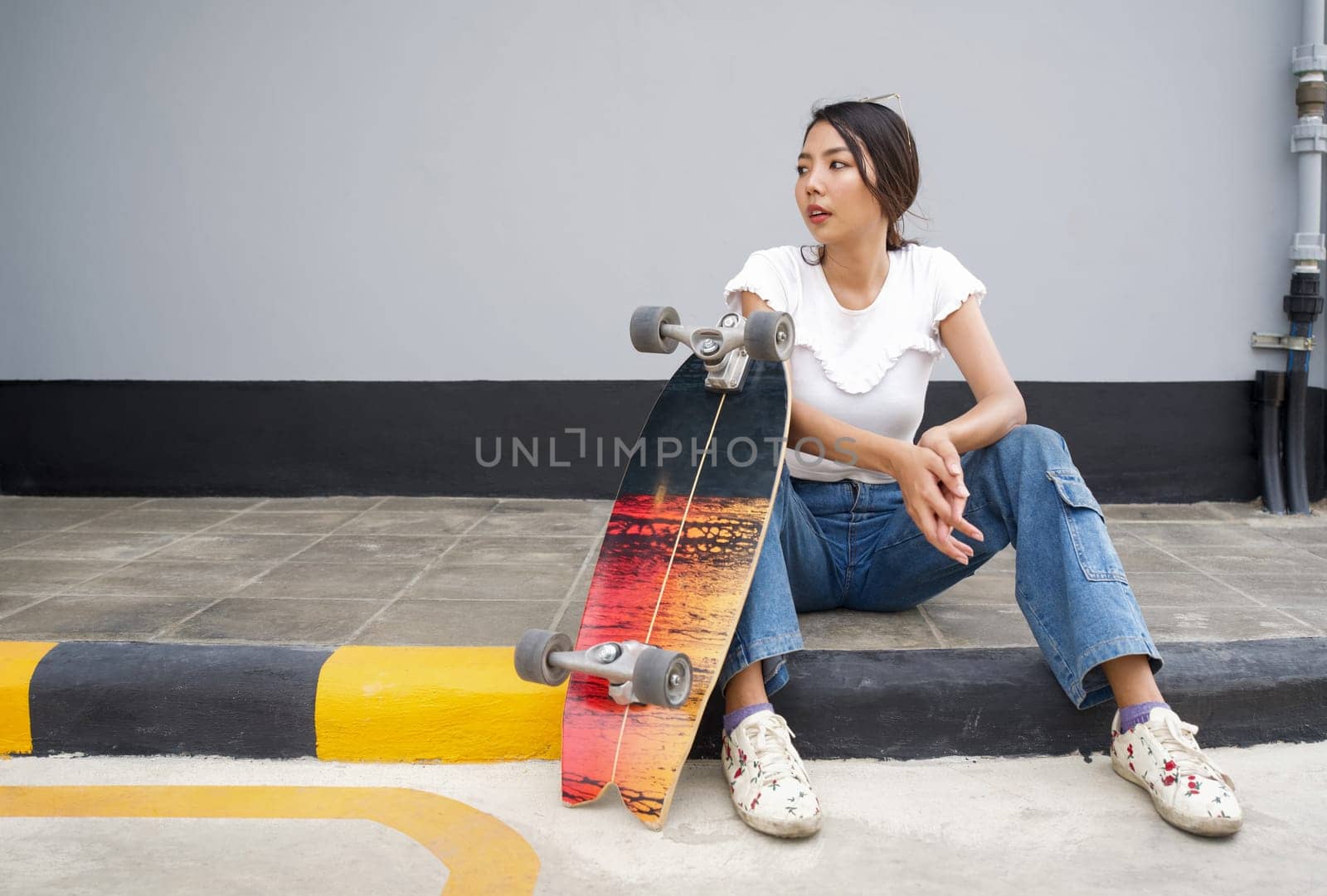 Young asian woman with sunglasses on her head resting after skateboarding. The skateboard is placed on the edge of the pedestrian lane.