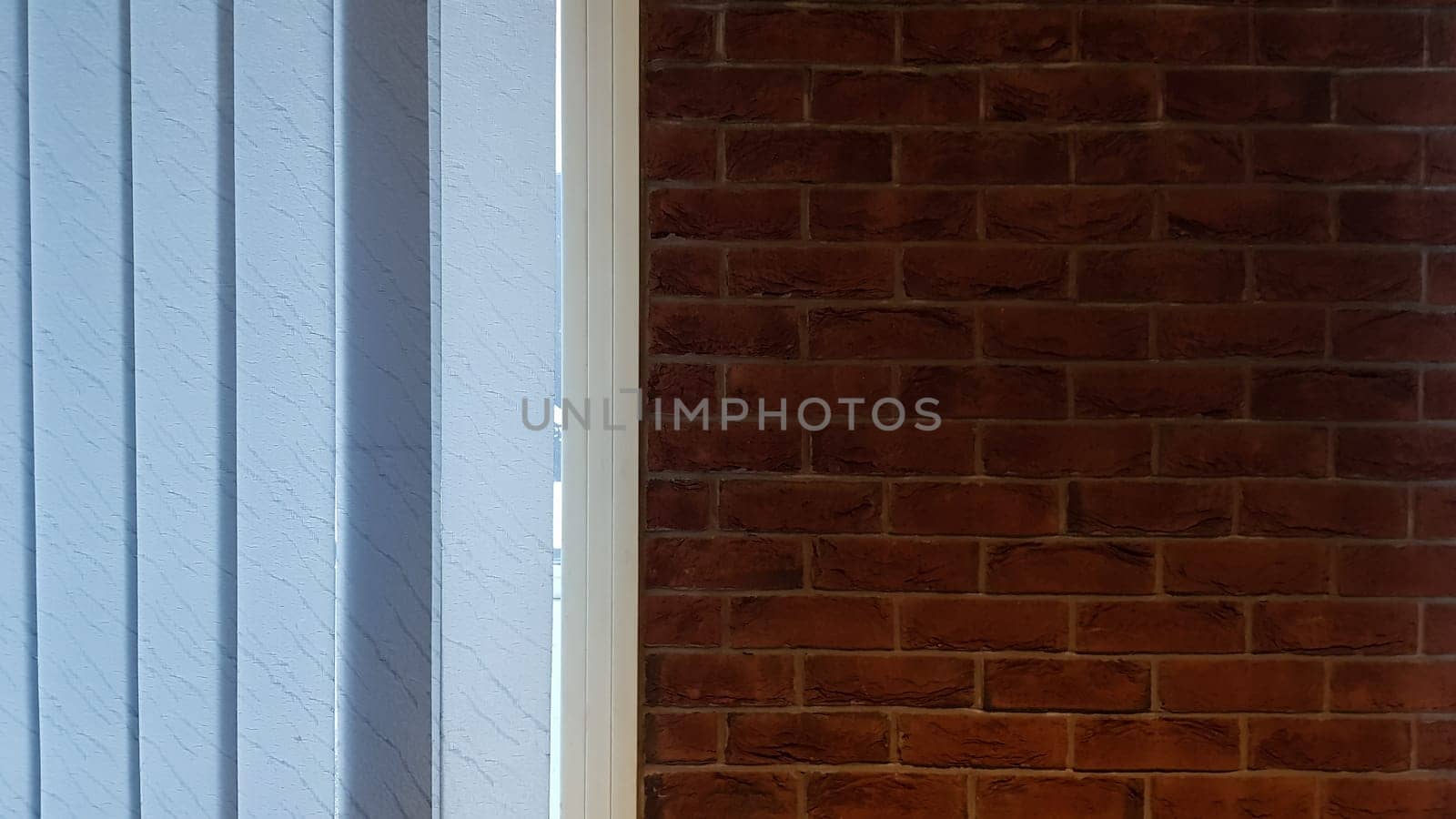 Large window with vertical white or gray blinds, loft, brick wall, industrial interior. Sunlight from a window on brick walls in a room or office