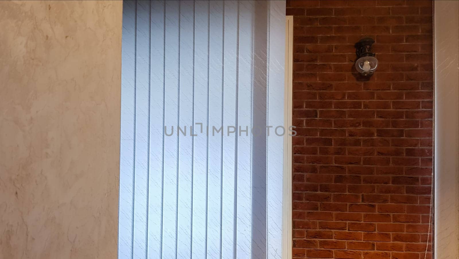 Large window with vertical white or gray blinds, loft, brick wall, industrial interior. Sunlight from a window on brick walls in a room or office