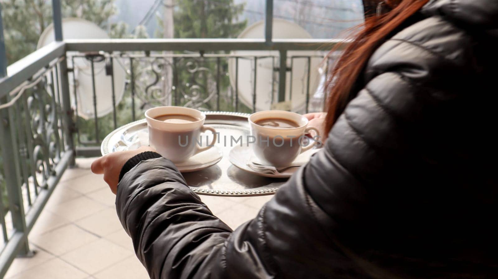 two cups of coffee on a tray in the hands of a girl. A woman with cups in her hands enters the hotel's balcony in the morning. Drink coffee with mountain and forest views