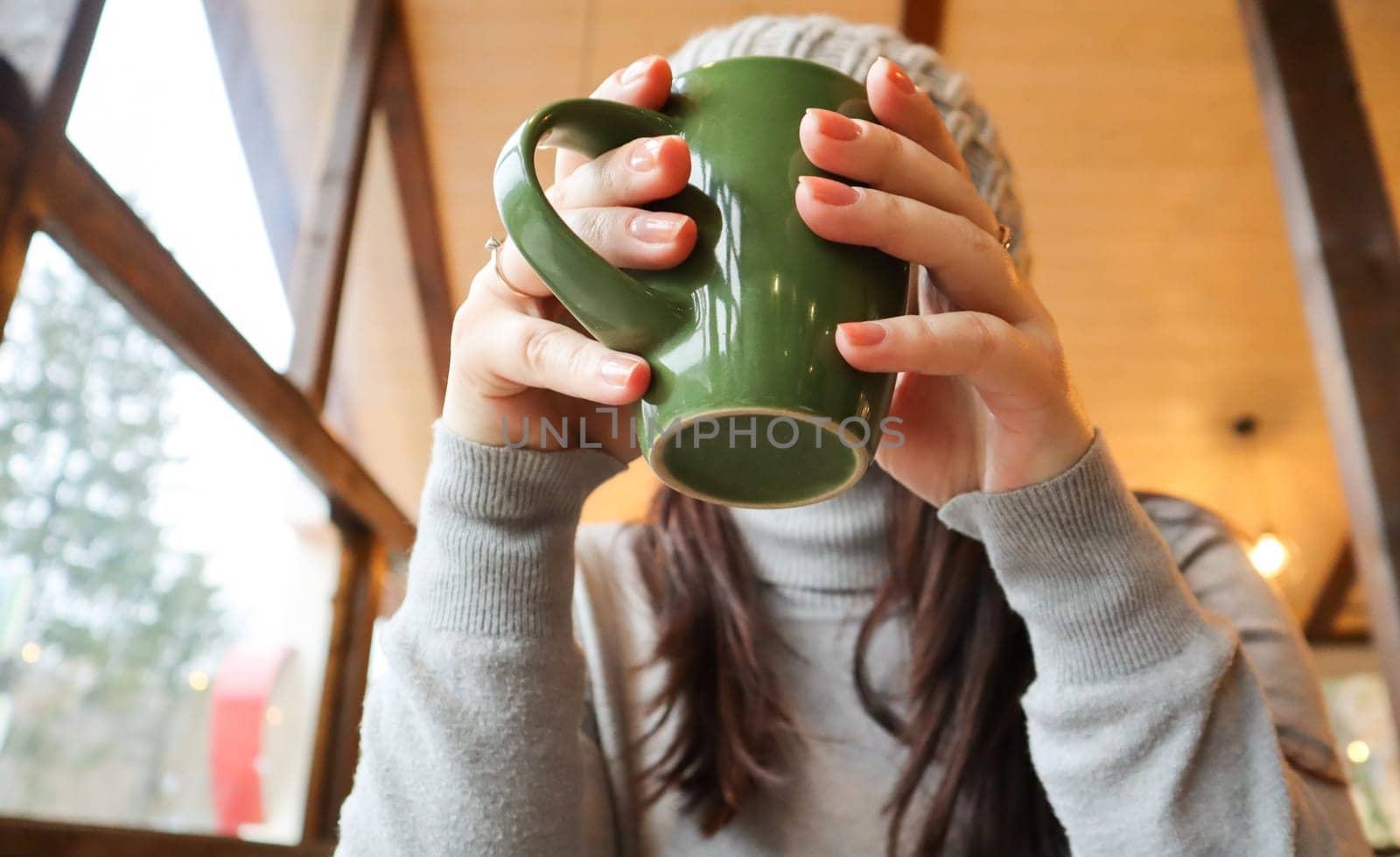 Close up of female hands with a drink mug. A girl in a gray sweater holds a cup of coffee or cocoa in a cafe at the table and looks out the window. Hot drink. A cozy photo with a blurry background.