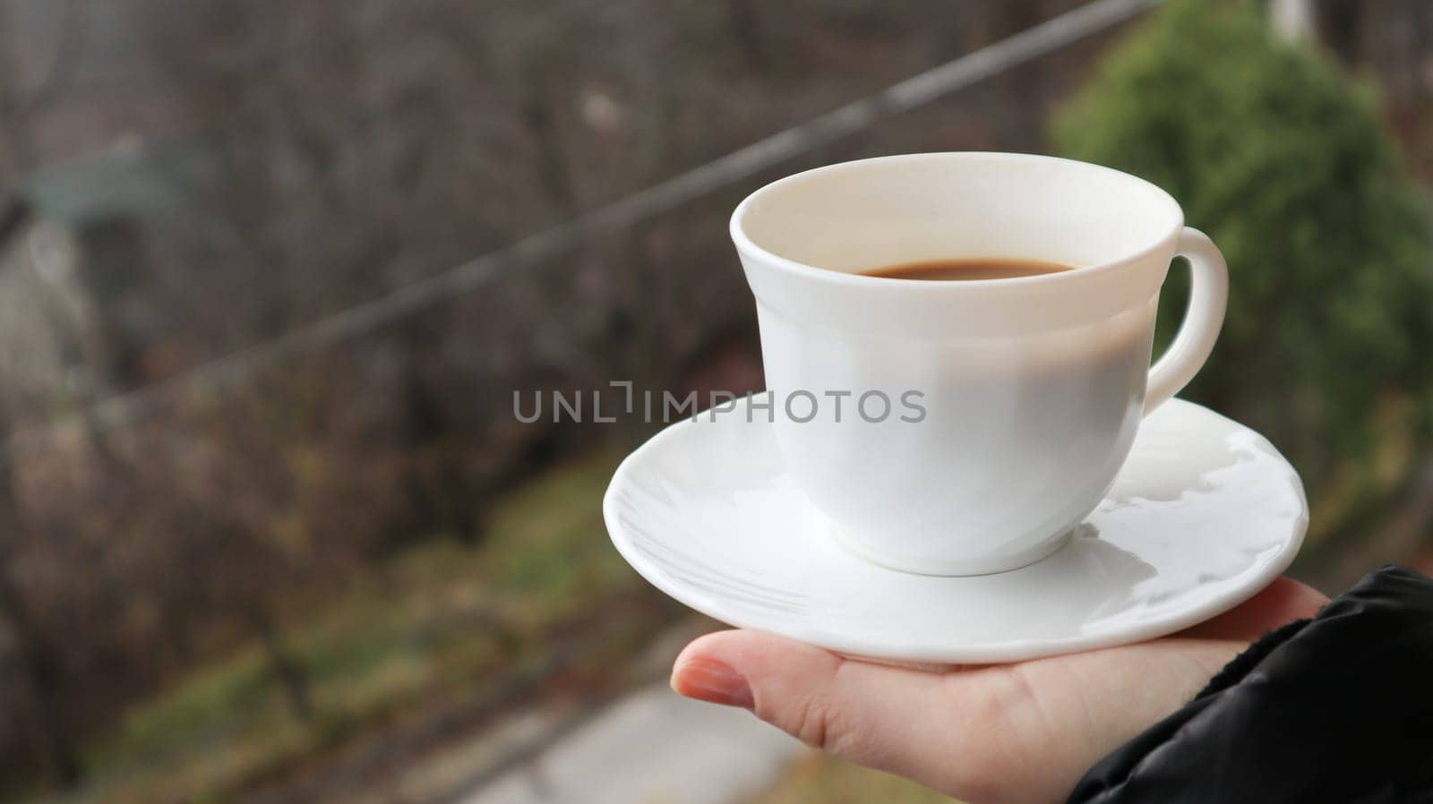 Girl hand holds a cup of white coffee and has a blurred green nature background. Mug in a female hand on a background of mountains