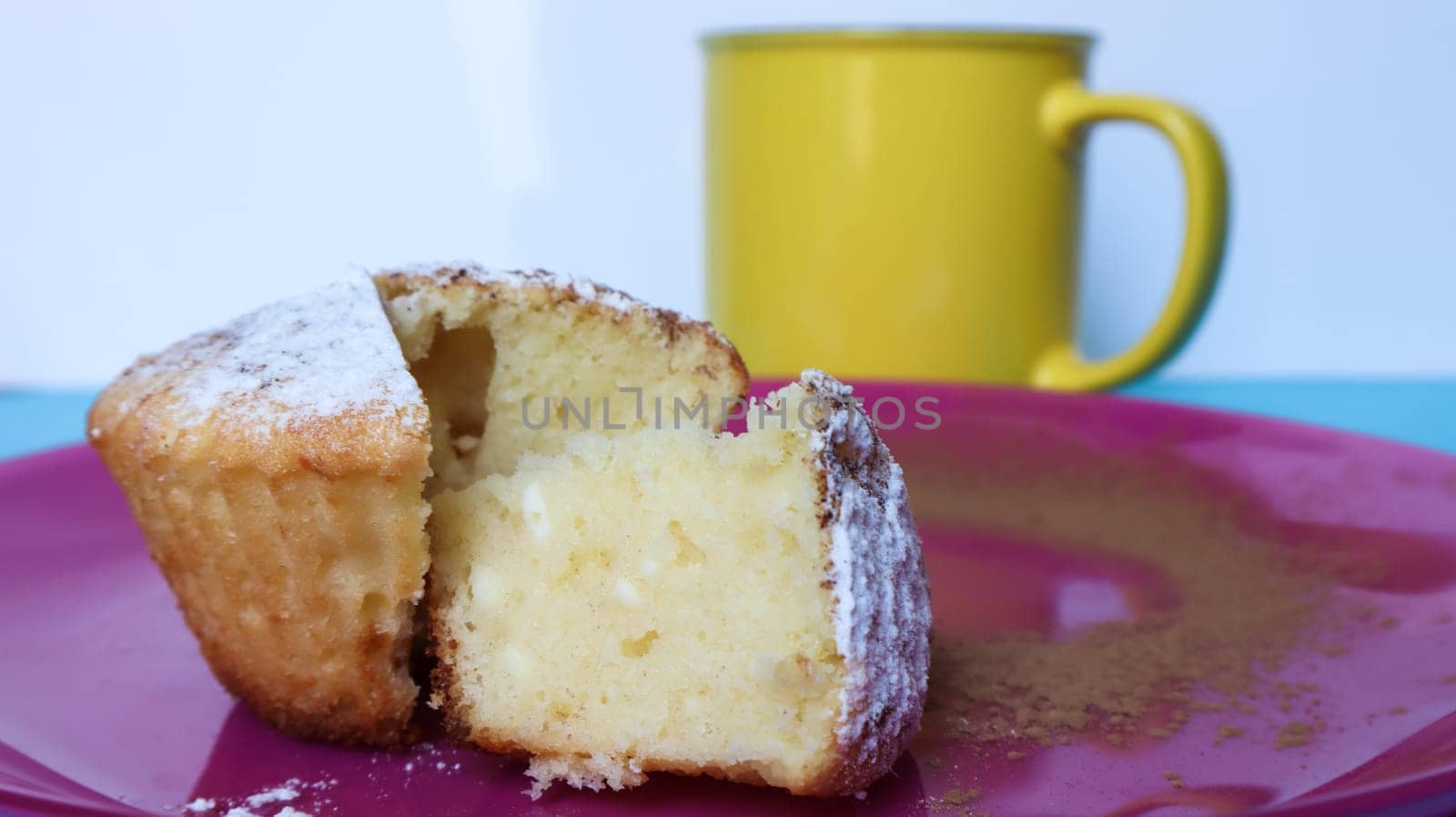 Side view of one cottage cheese sprinkled with icing sugar on a pink plate next to a yellow cup of coffee or tea. Dessert, a small cupcake. white baked cookies with an airy texture. Copy space.