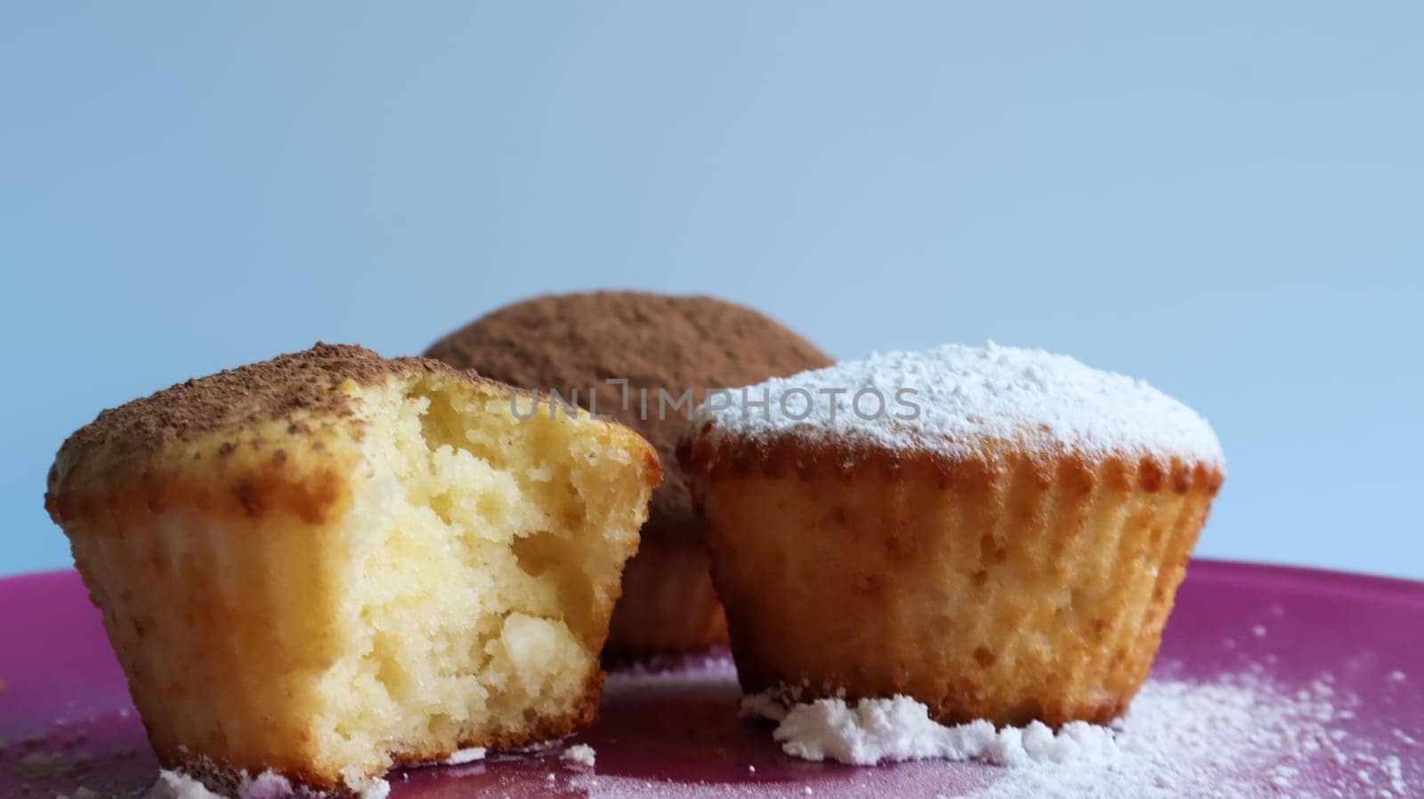 three curd cakes sprinkled with chocolate and icing sugar, one of them bitten, on a pink plate, on a blue background close-up. Dessert, a small cupcake. white baked cookies with an airy texture