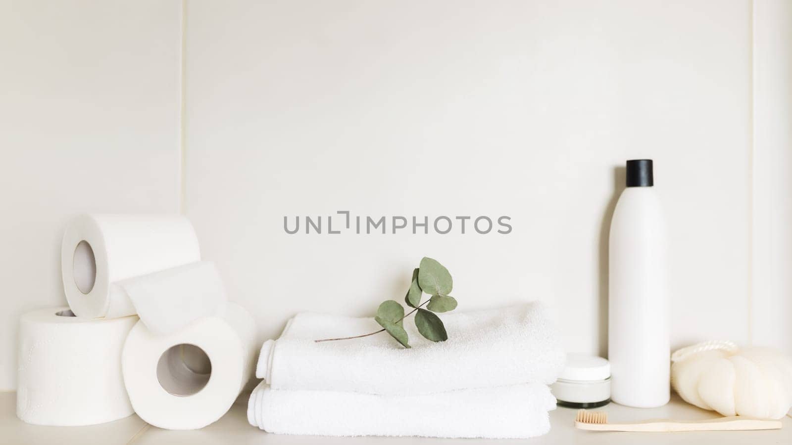 Washbasin in white bathroom, shelf with bath accessories. Hotel cleaning concept. Household concept. Washcloth, shampoo bottle, body cream, white toilet paper rolls, eucalyptus leaf, toothbrush