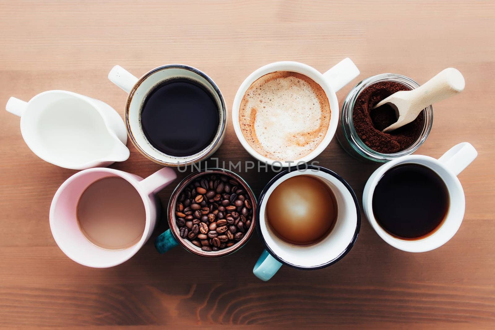 Multiple coffee cups and multicolored mugs with americano, espresso, latte and cappuccino, milk jar, roasted coffee beans in glass bottle, wooden spoon and ground coffee in jar on wooden background
