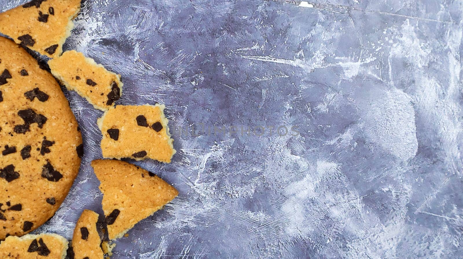 One soft freshly baked chocolate chip cookie with crumbs and chunks on a gray marble kitchen countertop. American traditional pastry, dessert. Delicious sweet food. Flat lay. Copy space.