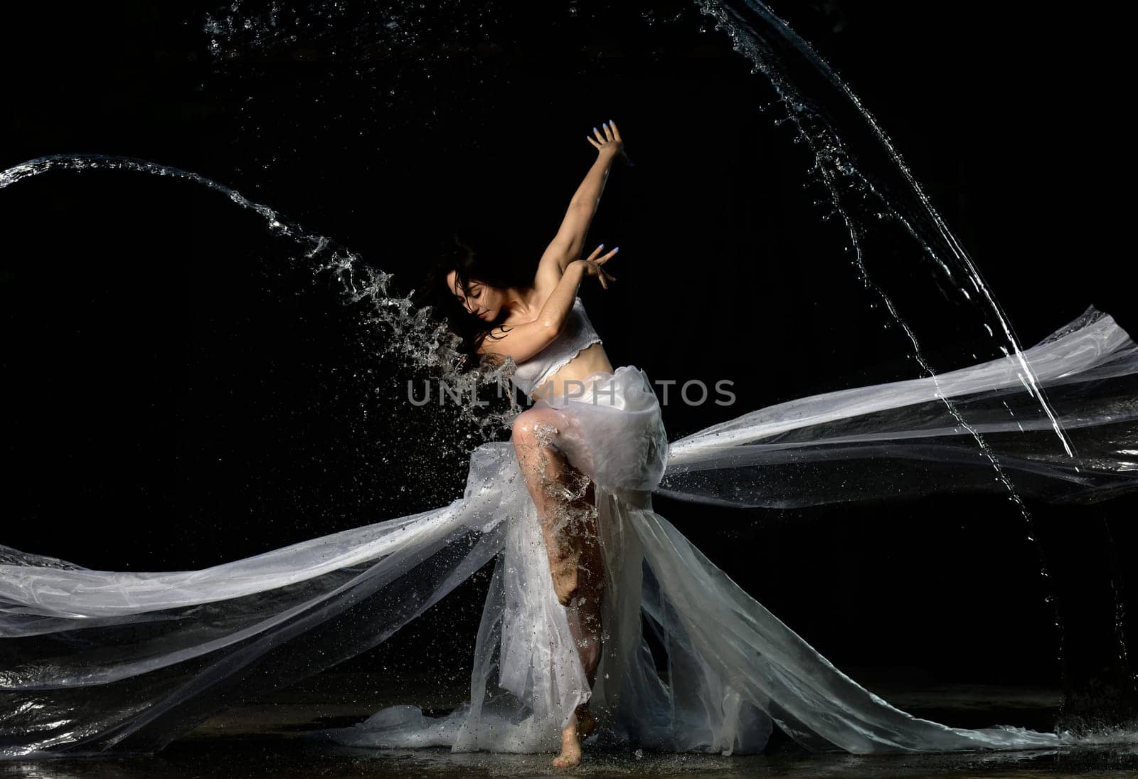 young beautiful woman of Caucasian appearance with long hair dances in drops of water on a black background. Jumping and swinging arms