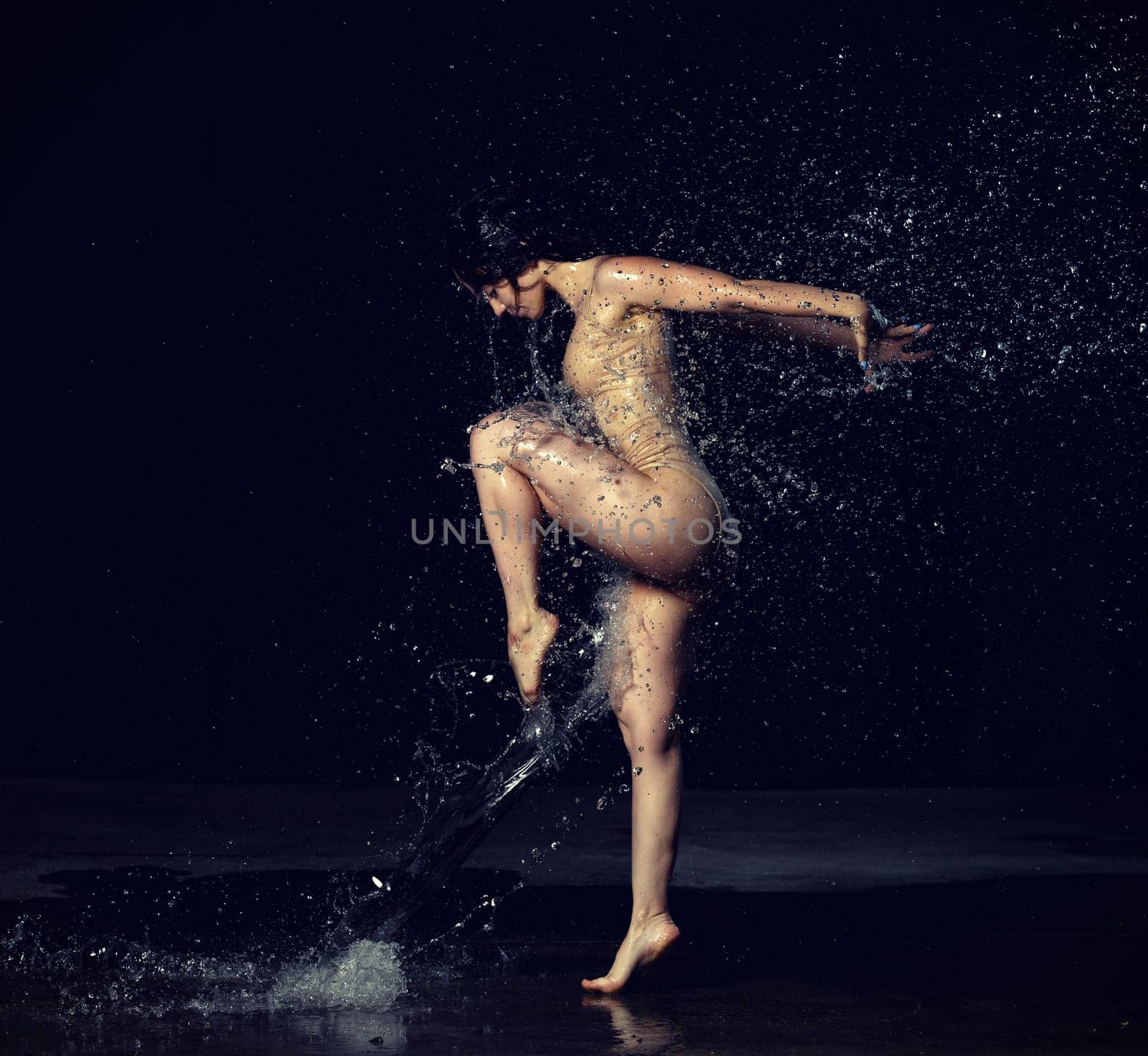 young beautiful woman of Caucasian appearance with long hair dances in drops of water on a black background. Jumping and swinging arms, dressed in a beige bodysuit