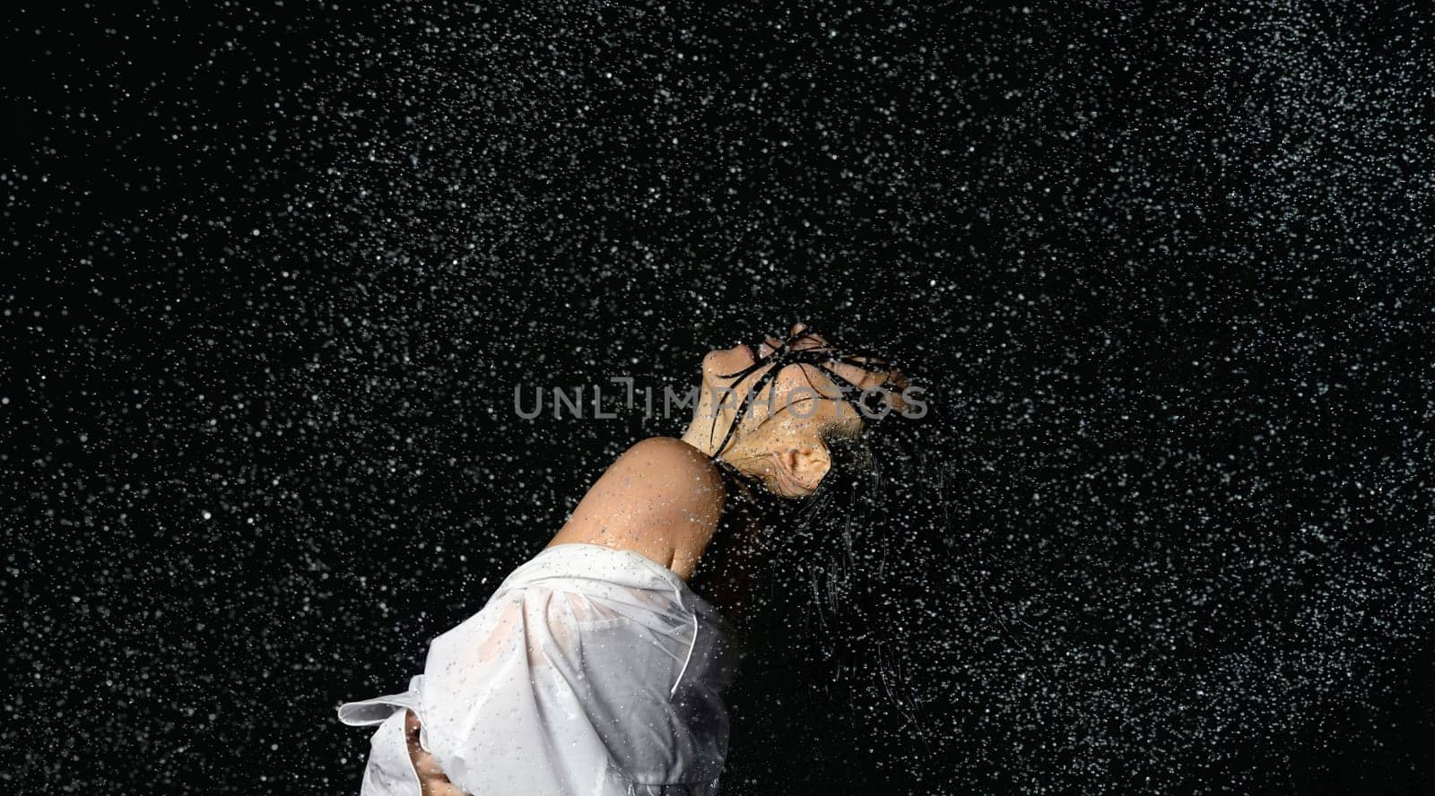 beautiful woman of Caucasian appearance with black hair dances in drops of water on a black background. Woman wearing white shirt