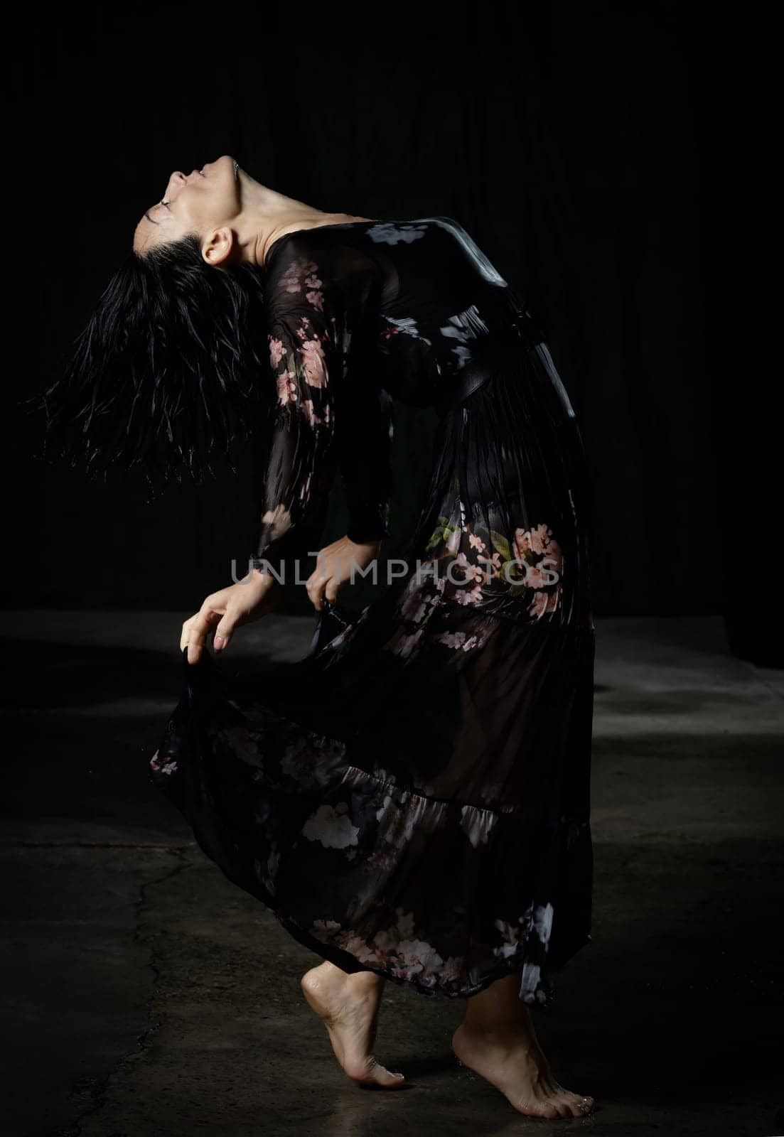 young woman with black hair dances in a black wet long chiffon dress on a dark background. The woman is bent back