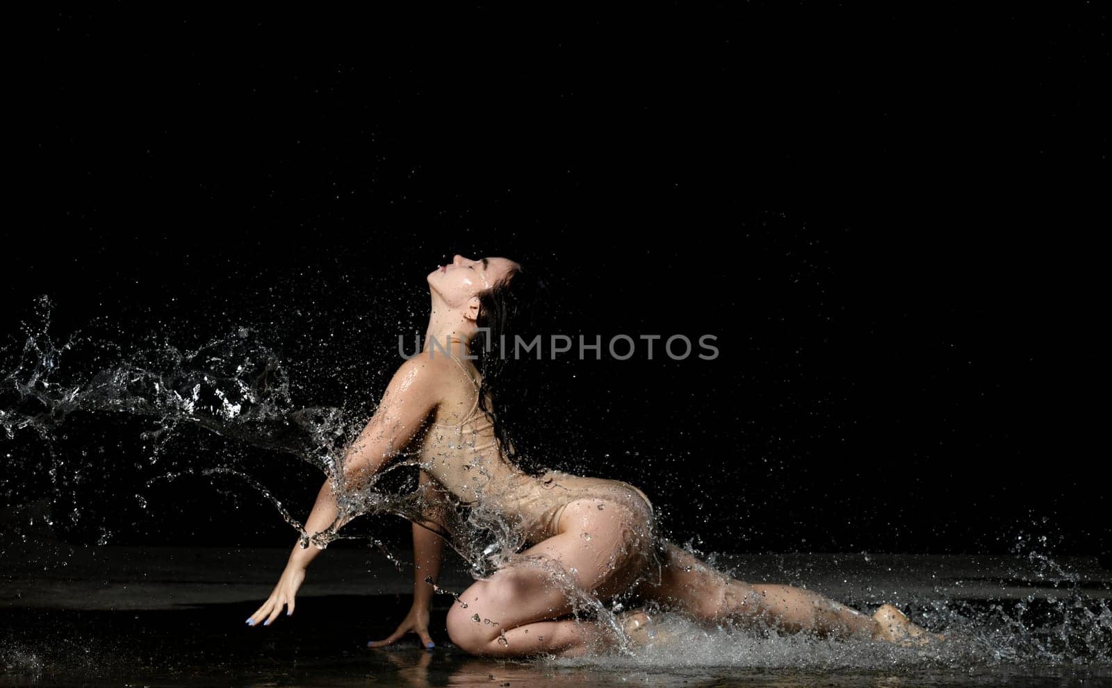 young woman with black long hair lies on the ground under raindrops on a black background. Woman dressed in beige bodysuit