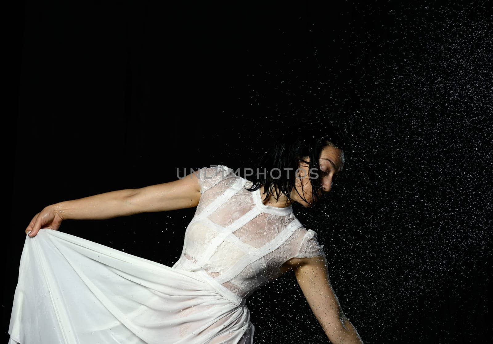 beautiful woman of Caucasian appearance with black hair dances in drops of water on a black background. The woman is wearing a white chiffon dress