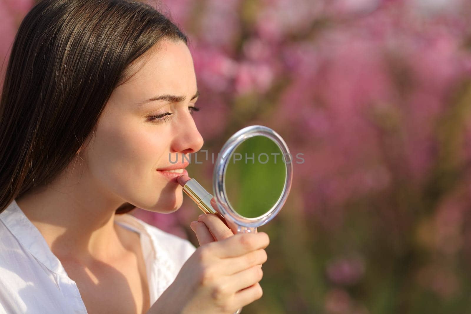 Beautiful concentrated woman painting lips using mirror in a pink field