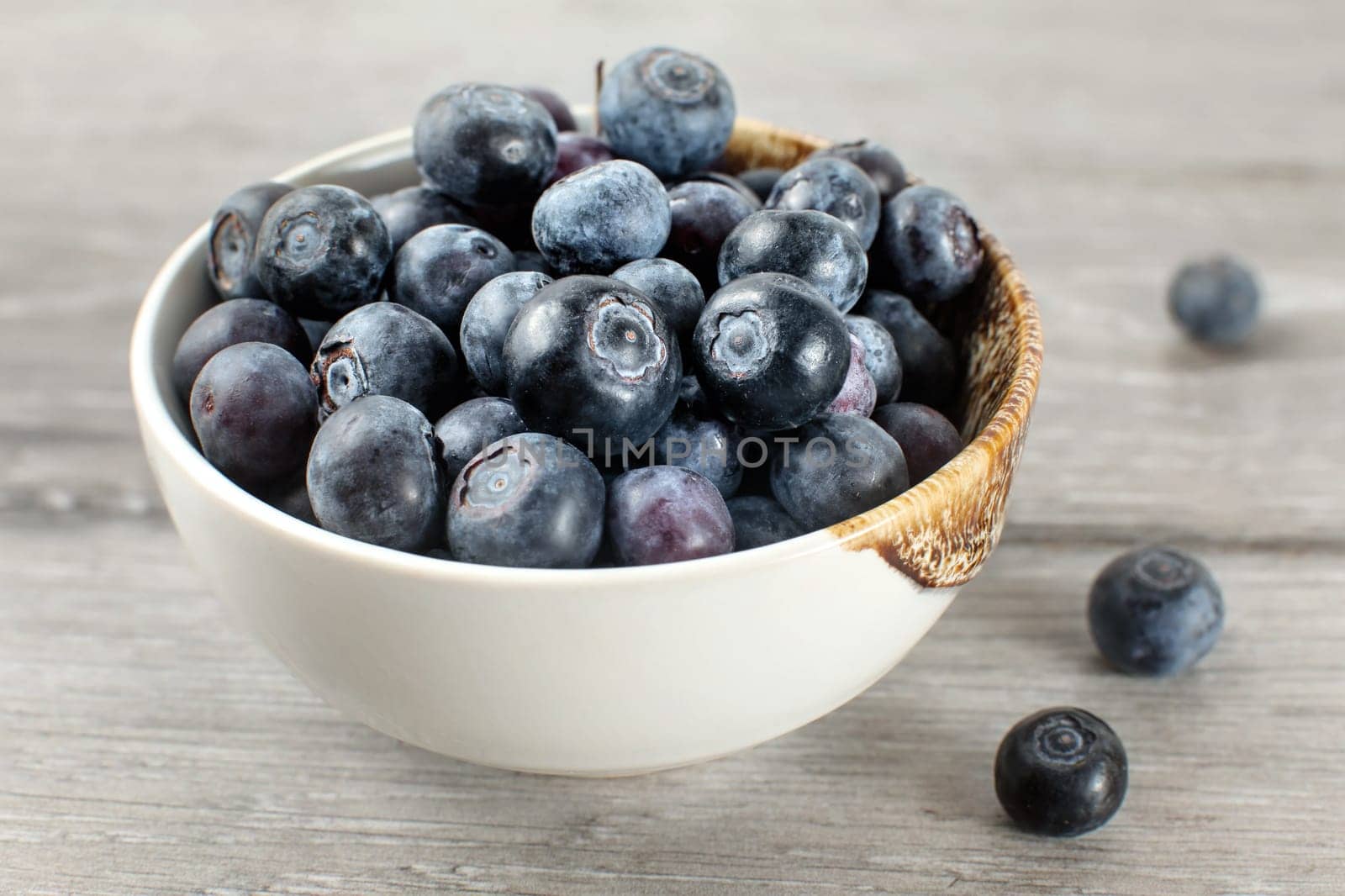 Small bowl full of blueberries, some of them spilled on gray wood desk.