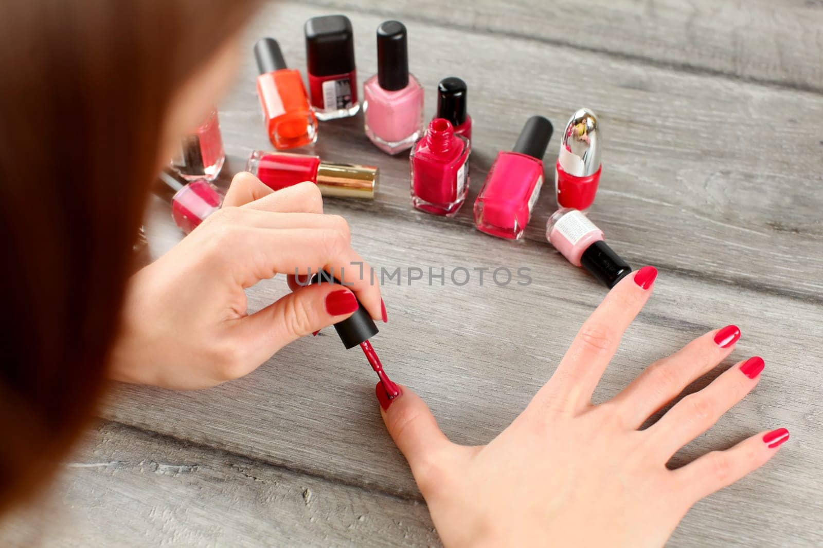 Top view on woman hands, applying red nail polish, with more bottles with varnish in background.