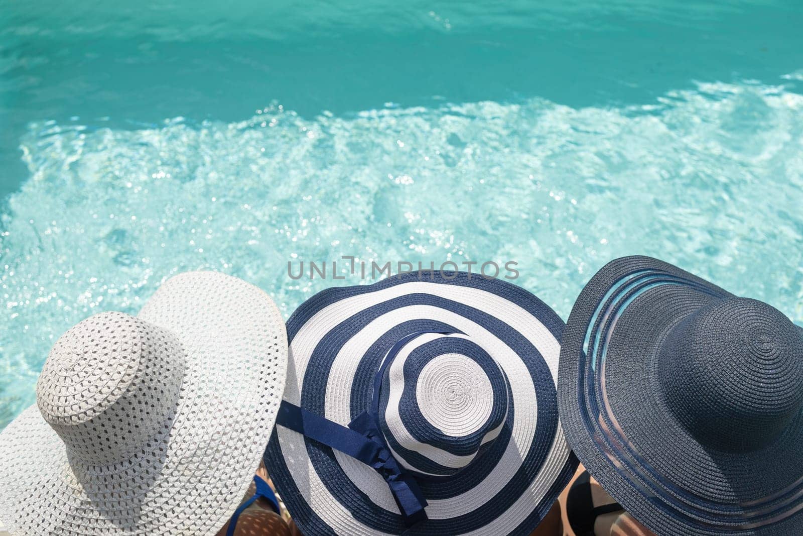 Top view of head of three women wearing straw hat sitting on the edge of the swimming pool. Horizontally.