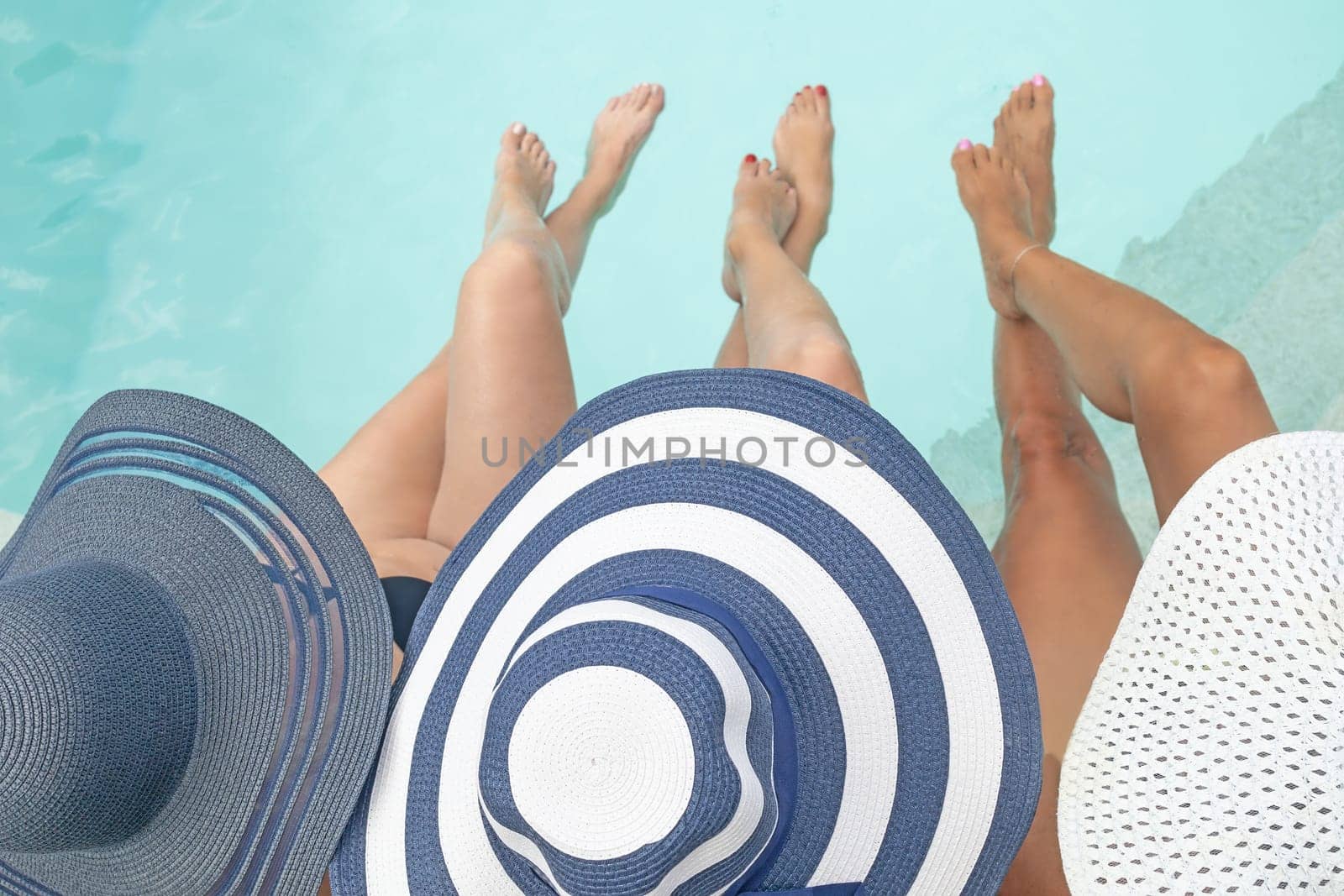 Top view of three women in straw hat relaxing  near a swimming pool with legs in water. Horizontally.