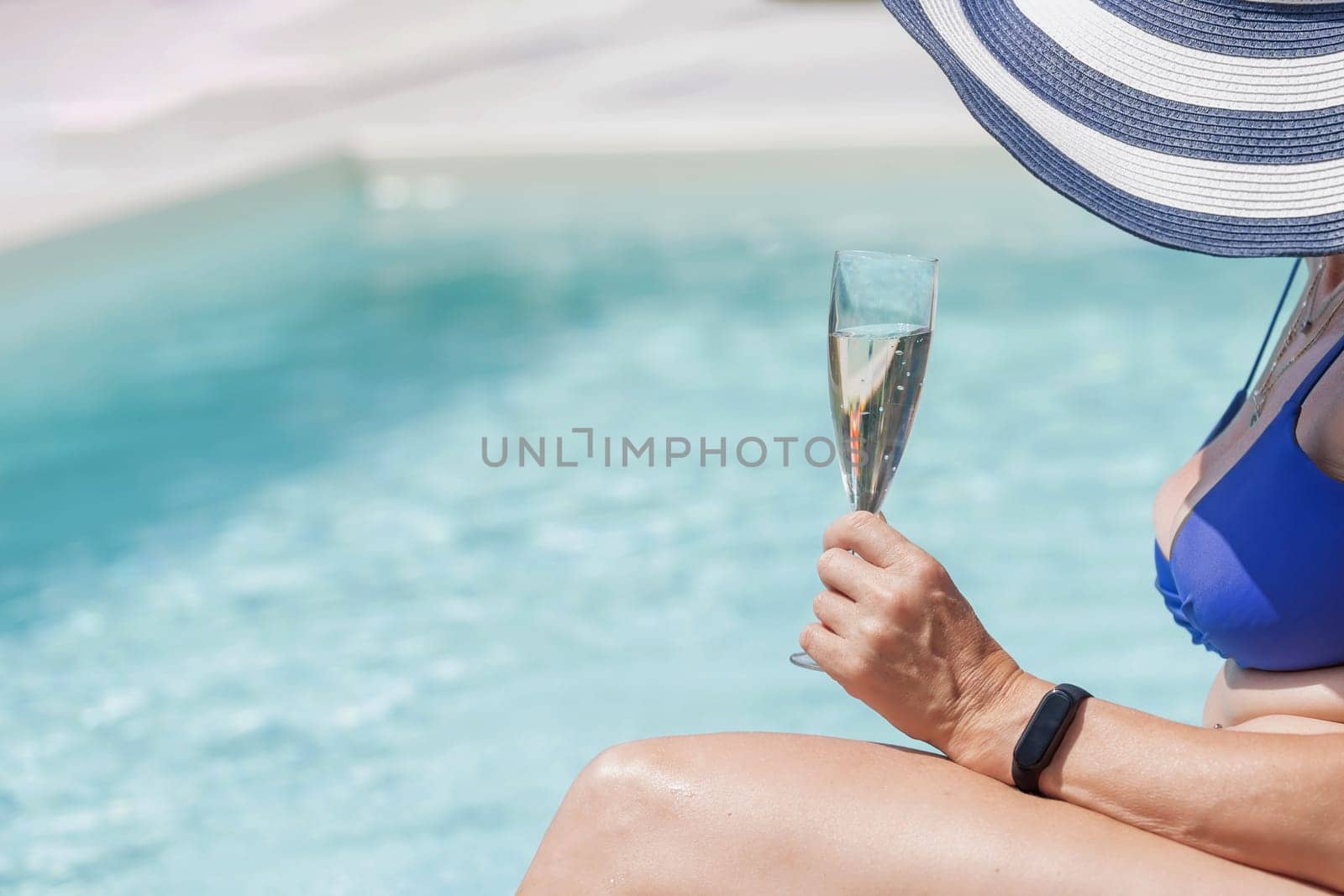 Middle aged female model in straw hat is holding glass of champagne sitting in the pool.
